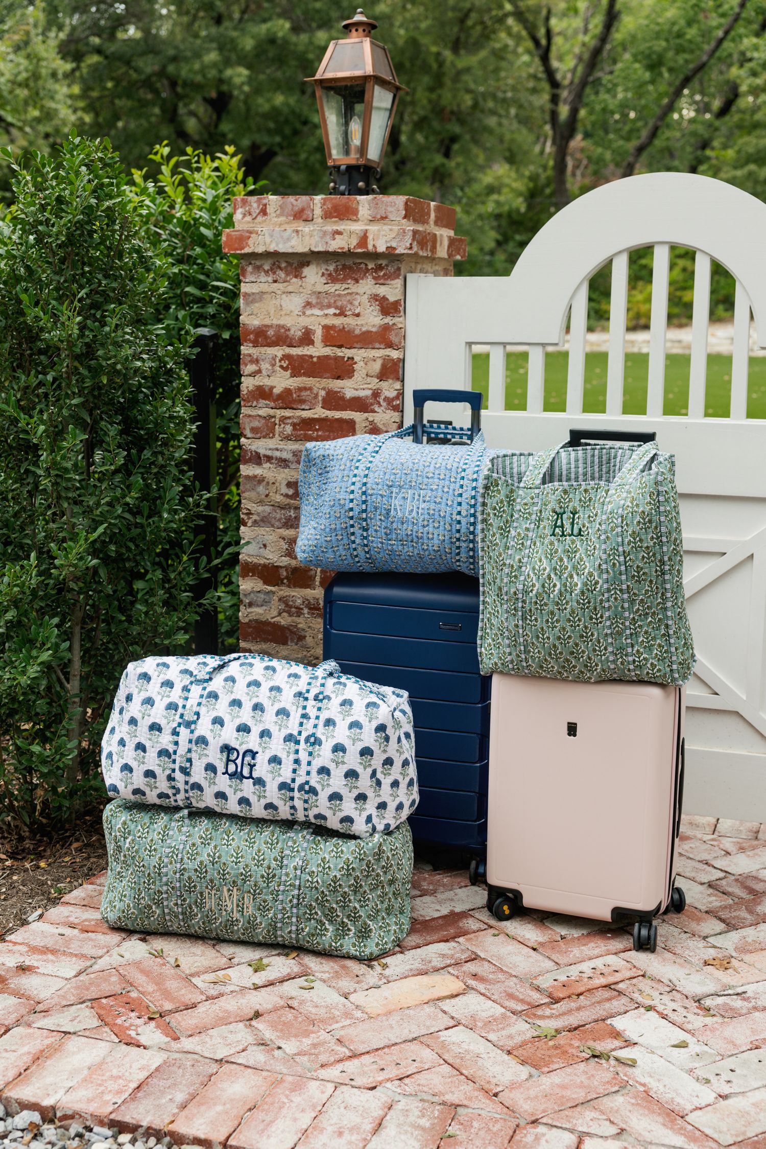 Monogrammed quilted travel bags and block print duffles stacked beside two suitcases at a white gate.