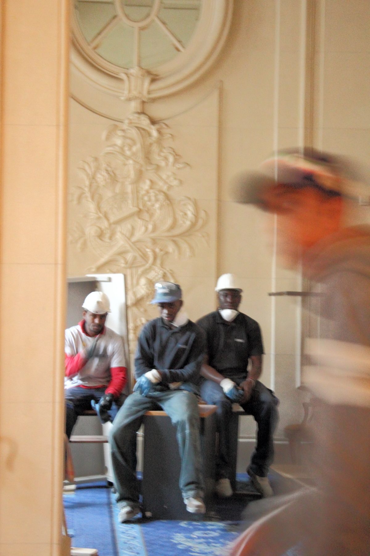 Three construction workers wearing hard hats sit on a bench inside an ornate decorated building.