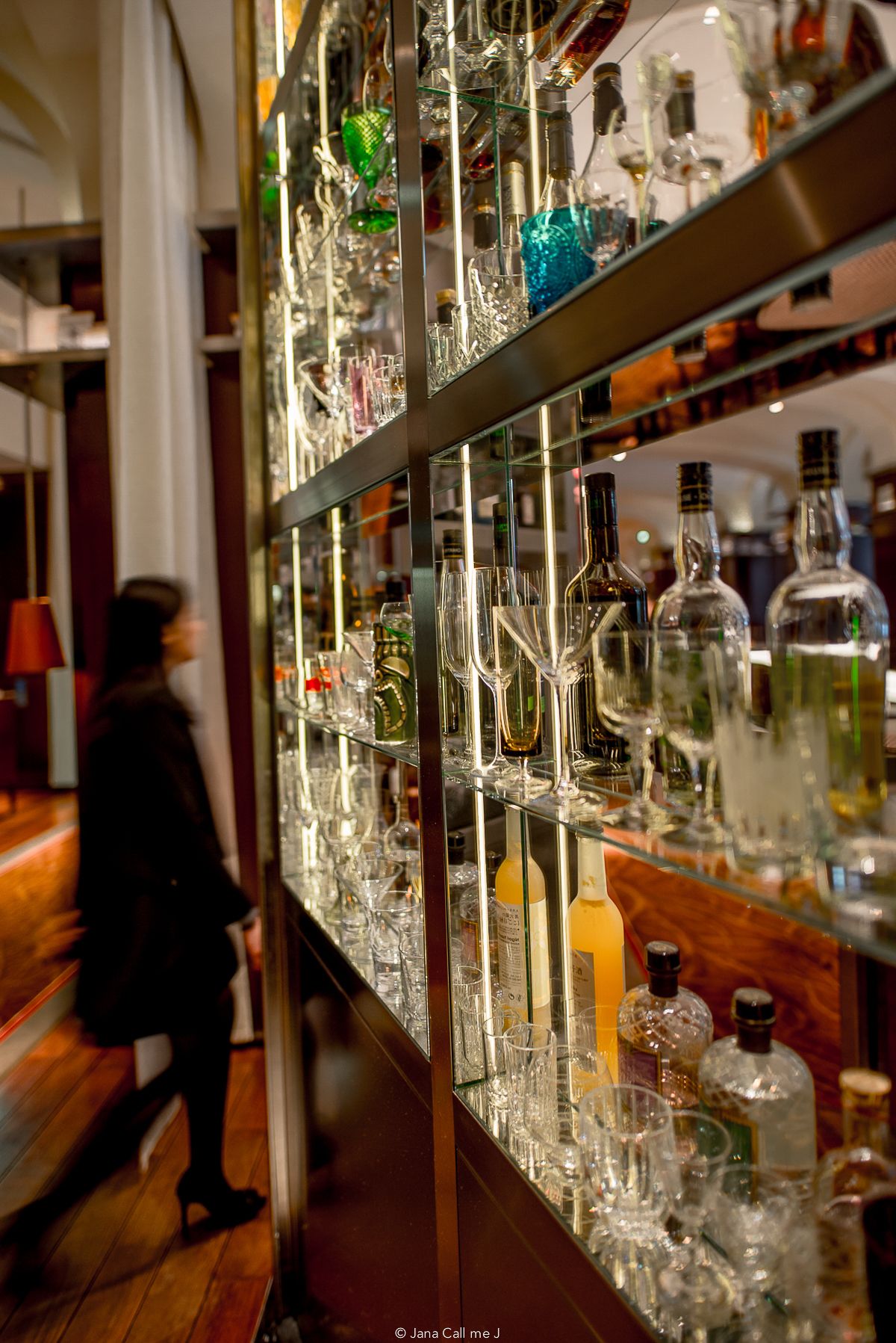 Elegant bar shelves stocked with premium liquor bottles and crystal glassware, with a blurred figure walking past.