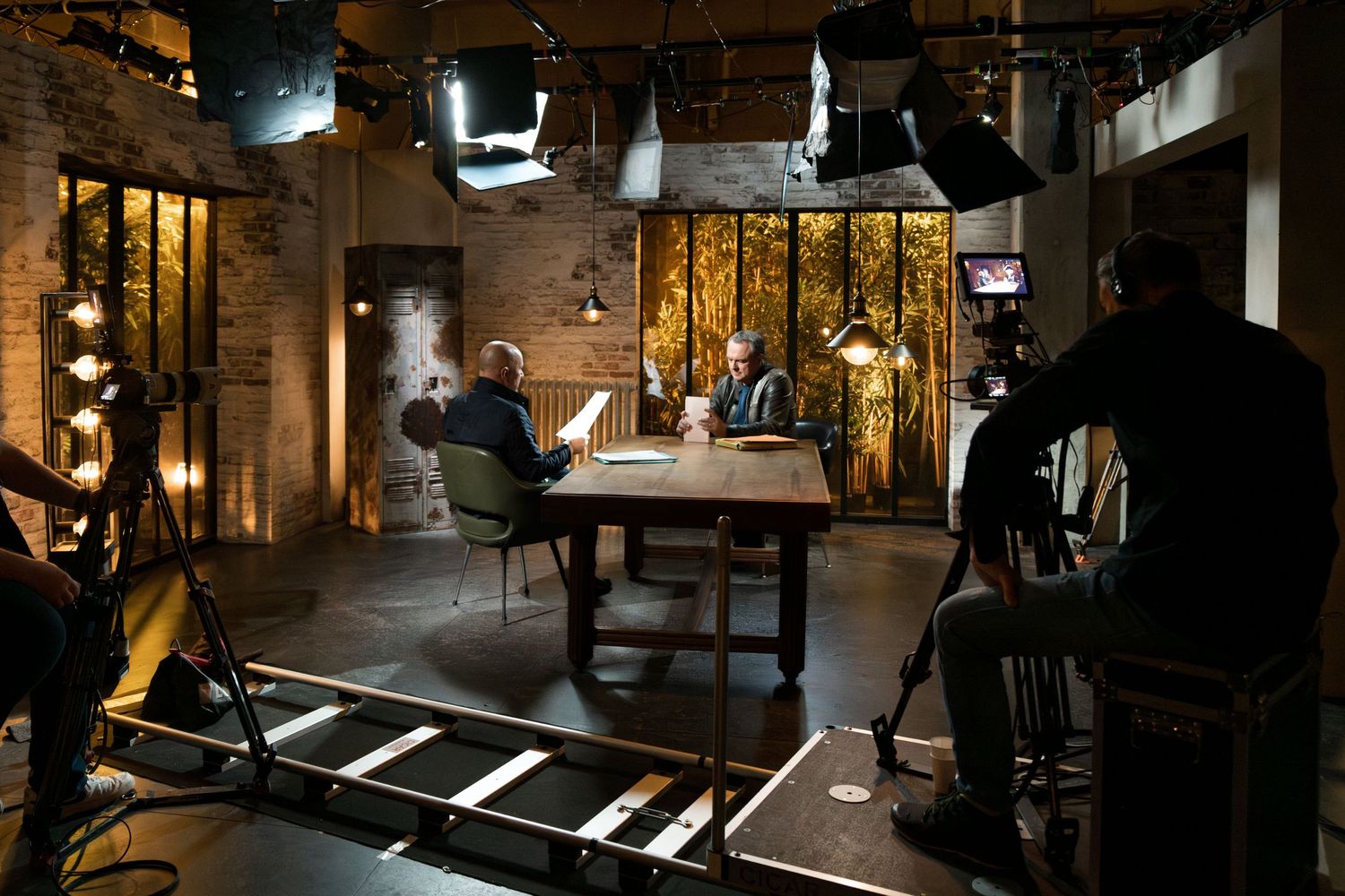 Two men filmed at a rustic TV studio set with professional lighting equipment and cameras.
