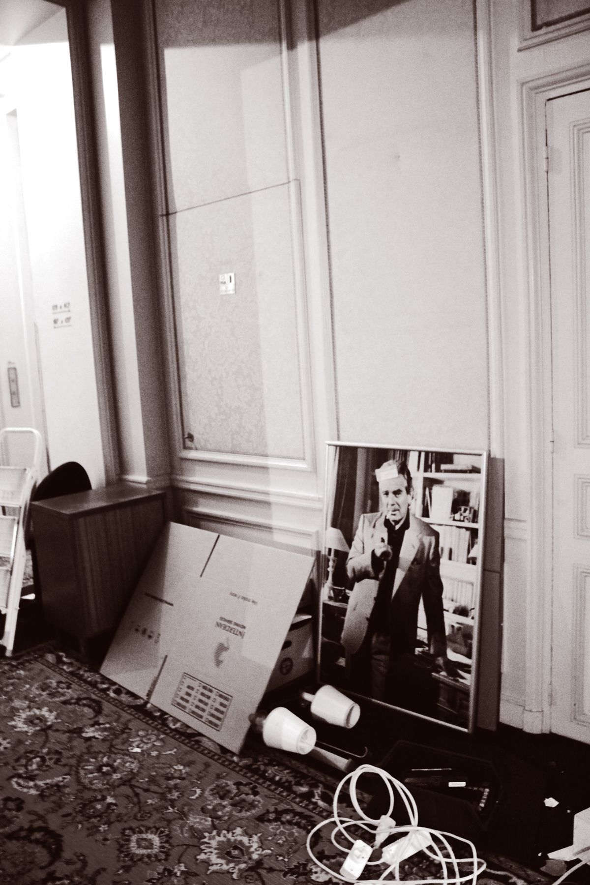 Black and white photo of a portrait print and cardboard boxes leaning against a wall in a hallway.