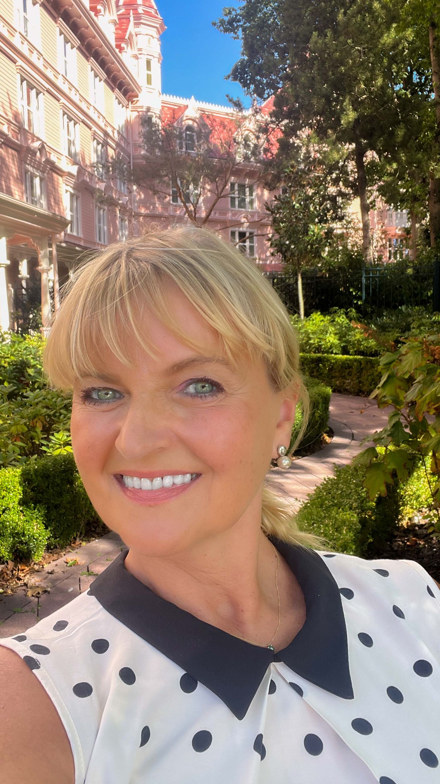 Smiling Jana Maillot in front of a Disneyland Hotel in Paris, red brick building surrounded by lush green gardens.