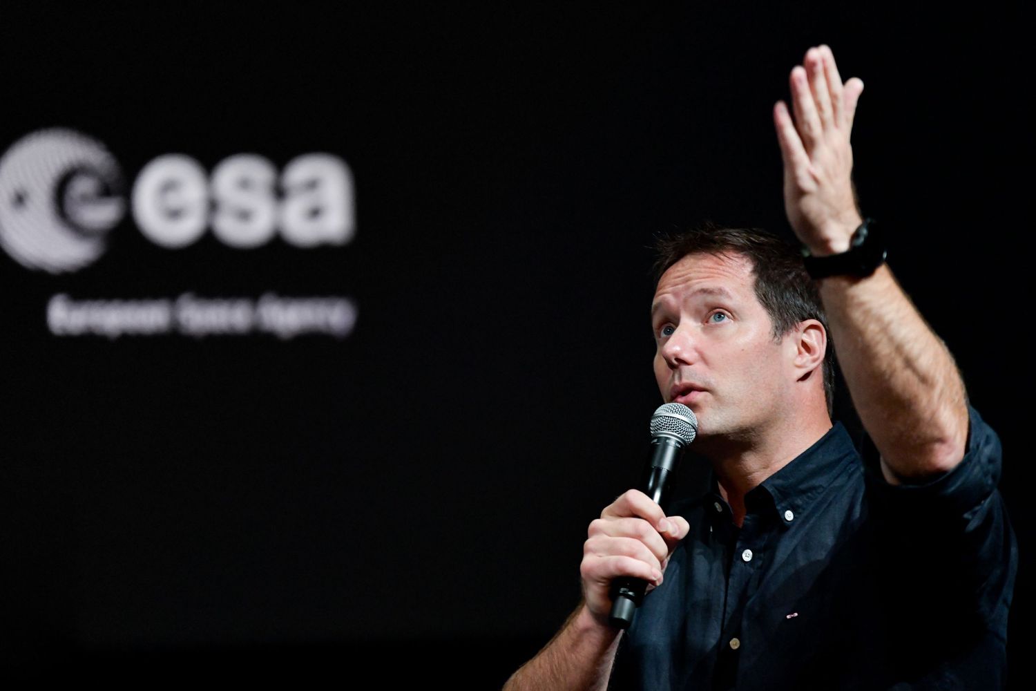 Thomas Pesquet speaking at ESA European Space Agency event, holding microphone and gesturing with raised hand.