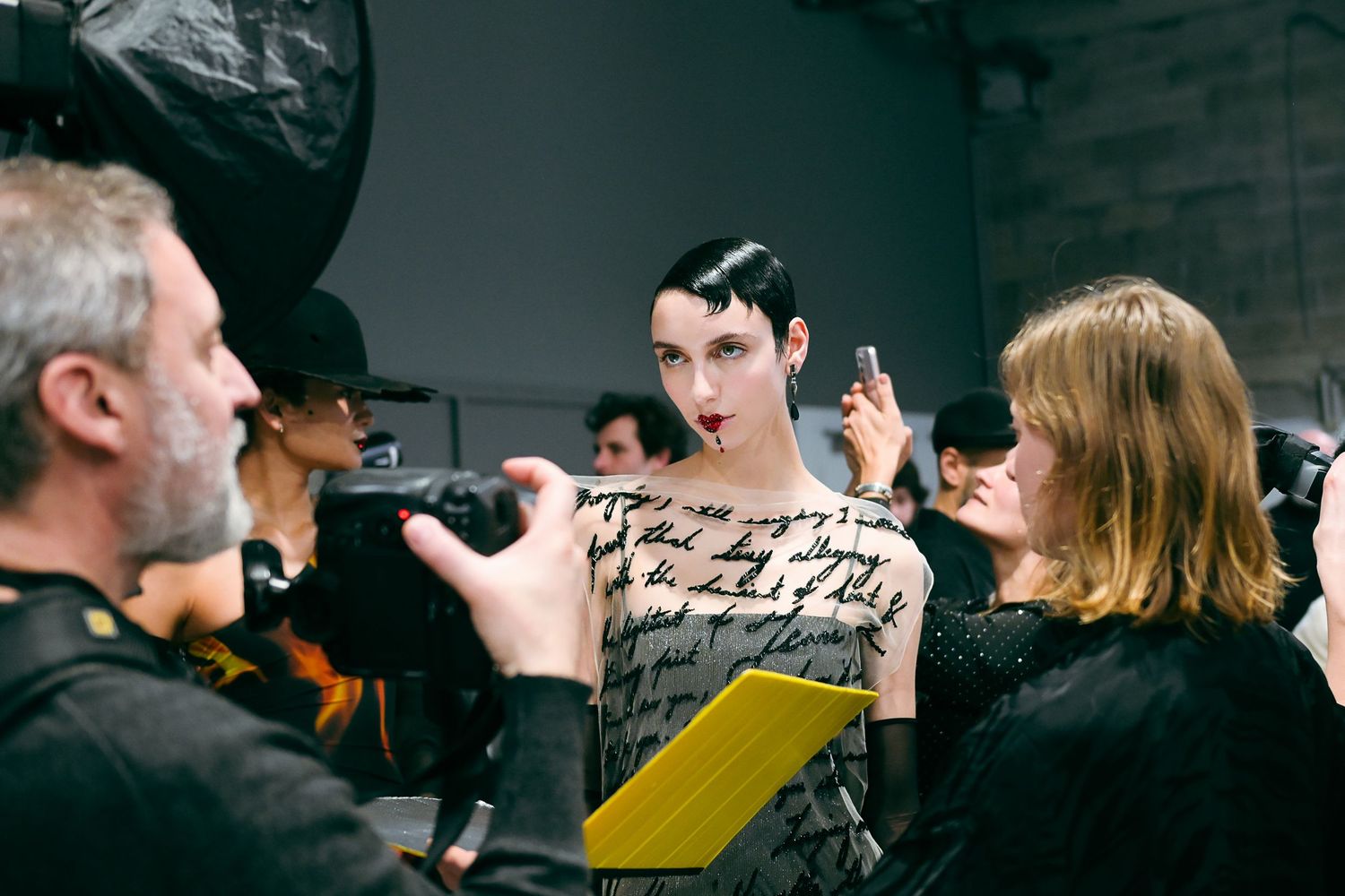 Model in patterned dress being photographed backstage at a fashion show of Robert Wun, with crew members.