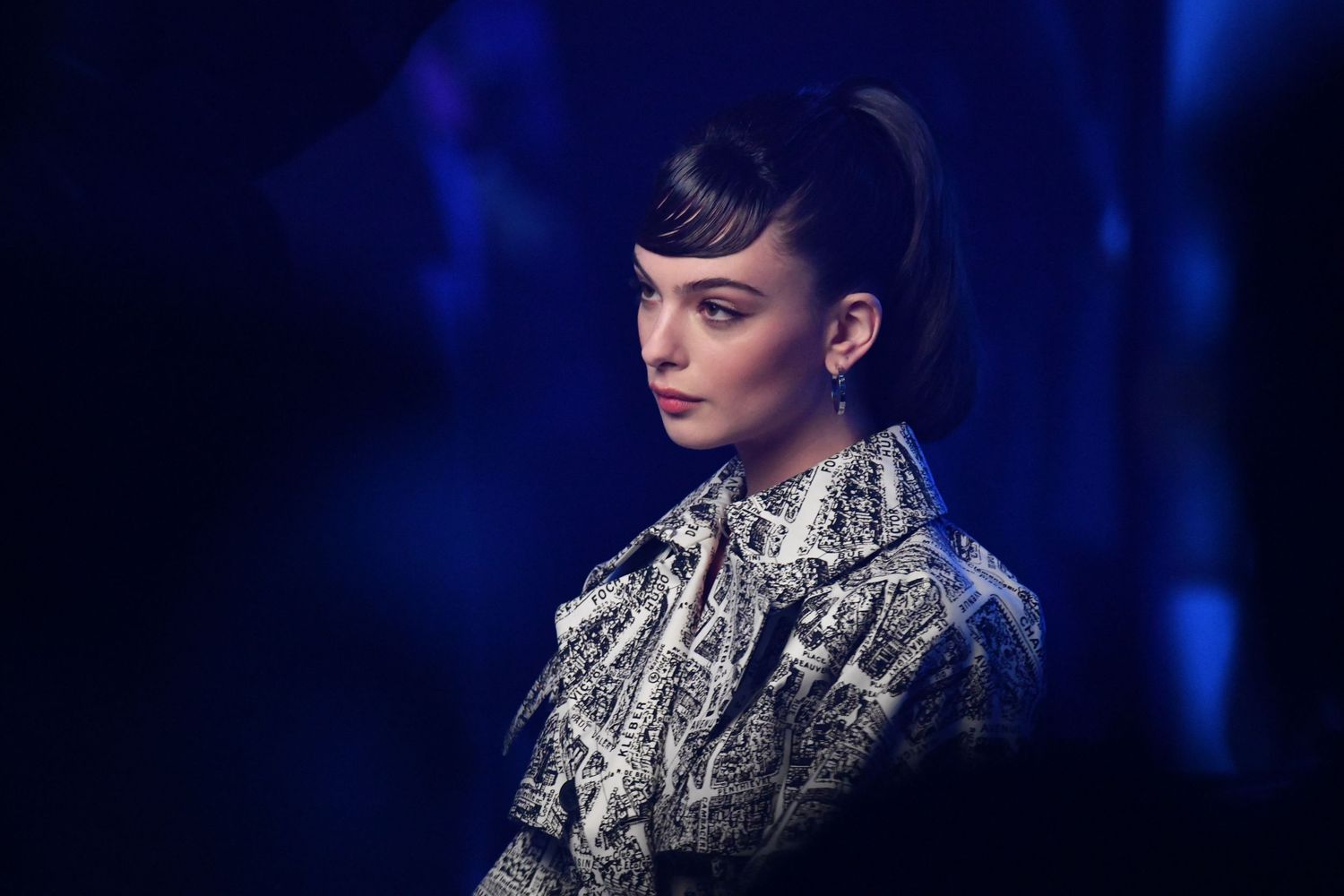 Deva Cassel wearing Dior with dark bangs in patterned jacket posing against a dramatic blue background.