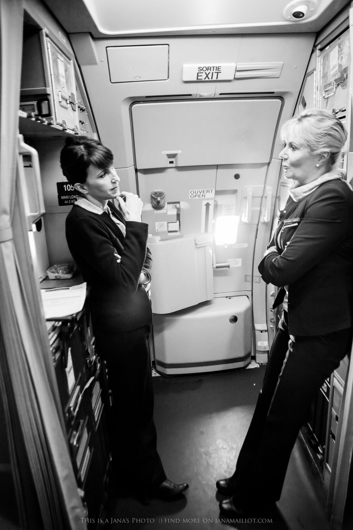 Black and white photo of two female flight attendants conversing near aircraft exit door in uniform.