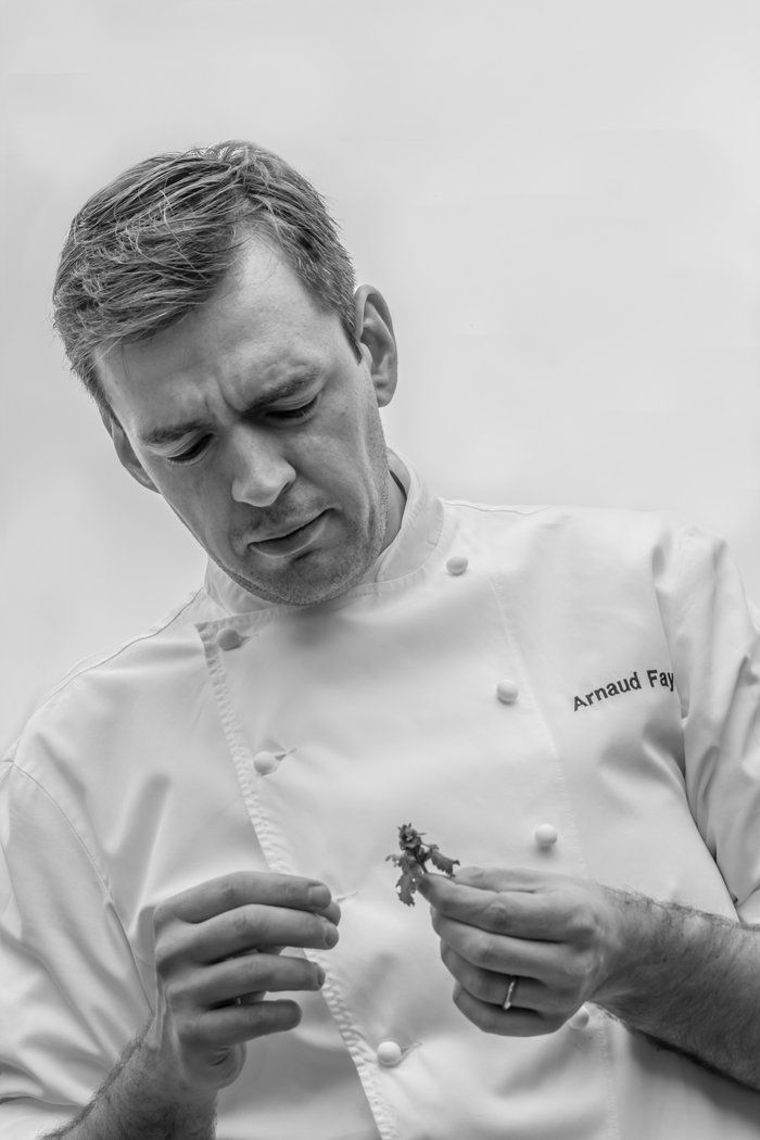 Black and white close-up of chef Arnaud Faye in white uniform carefully examining a small herb or garnish.