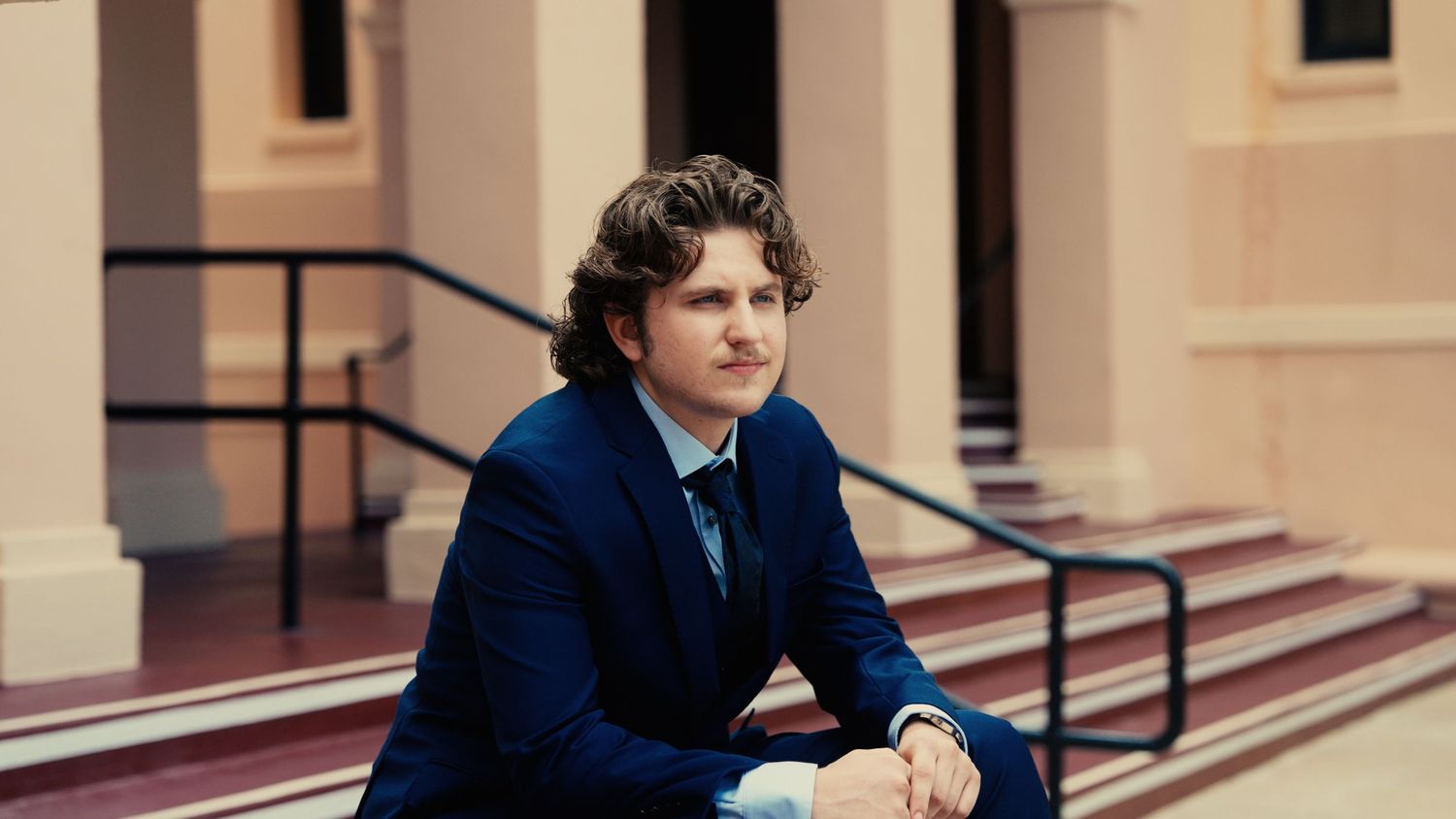 Young man in navy blue suit sitting on steps of classical building with pink columns.