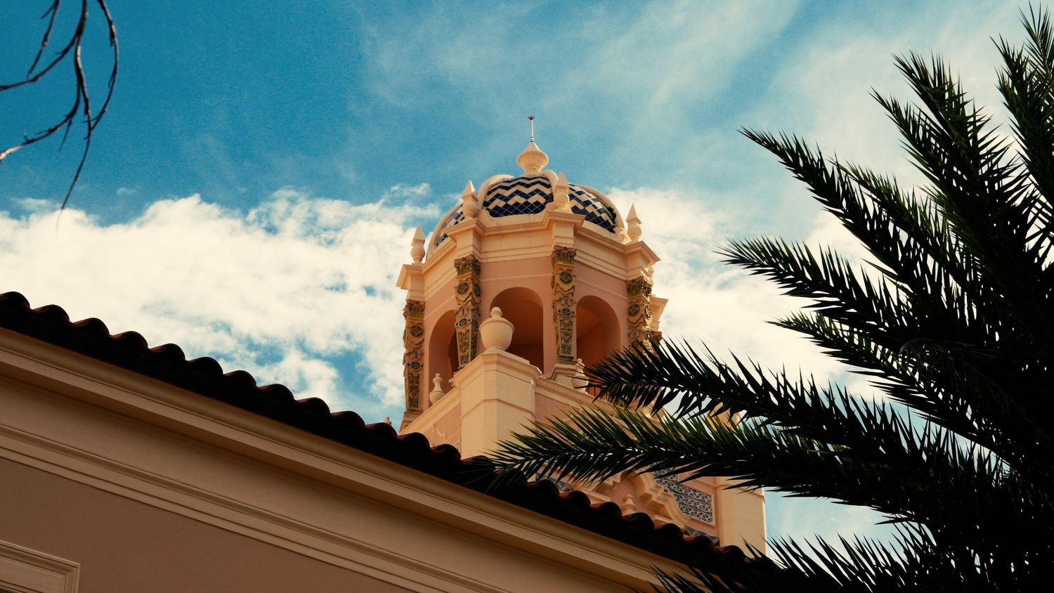 Elegant Spanish colonial church bell tower with palm tree and blue cloudy sky.