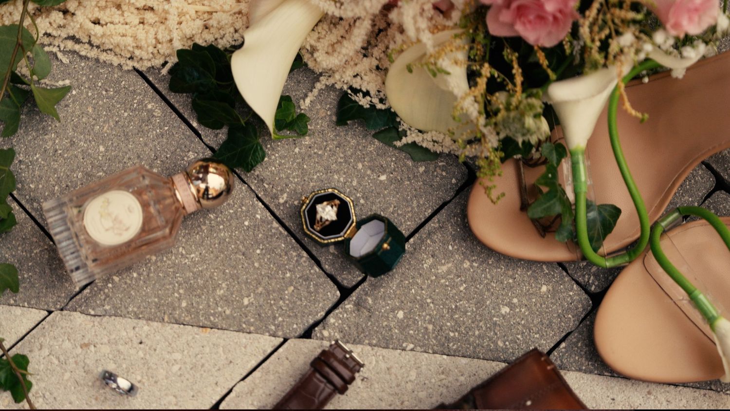 Flat lay of wedding accessories including perfume, rings, sunglasses, heels and flowers.