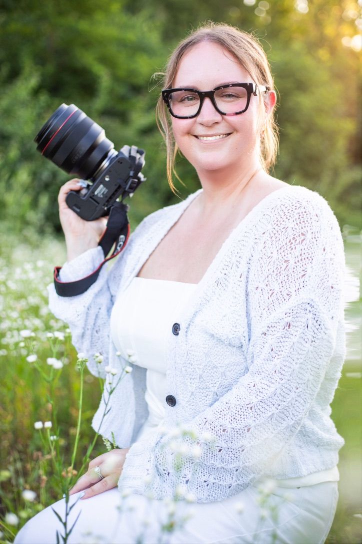 Dayton area photographer poses for photo. She is kneeling in a field of white wildflowers in Fairborn, OH and is holding her camera up.