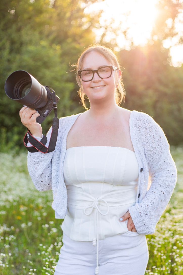 Photographer poses for photo in Dayton, Ohio. She is holding a camera and smiling. The sun is setting behind her, bathing her in a warm glow