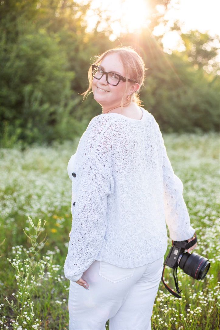 Dayton area photographer is walking a field of white wildflowers and looks back at the camera. She is smiling and the sun is glowing behind