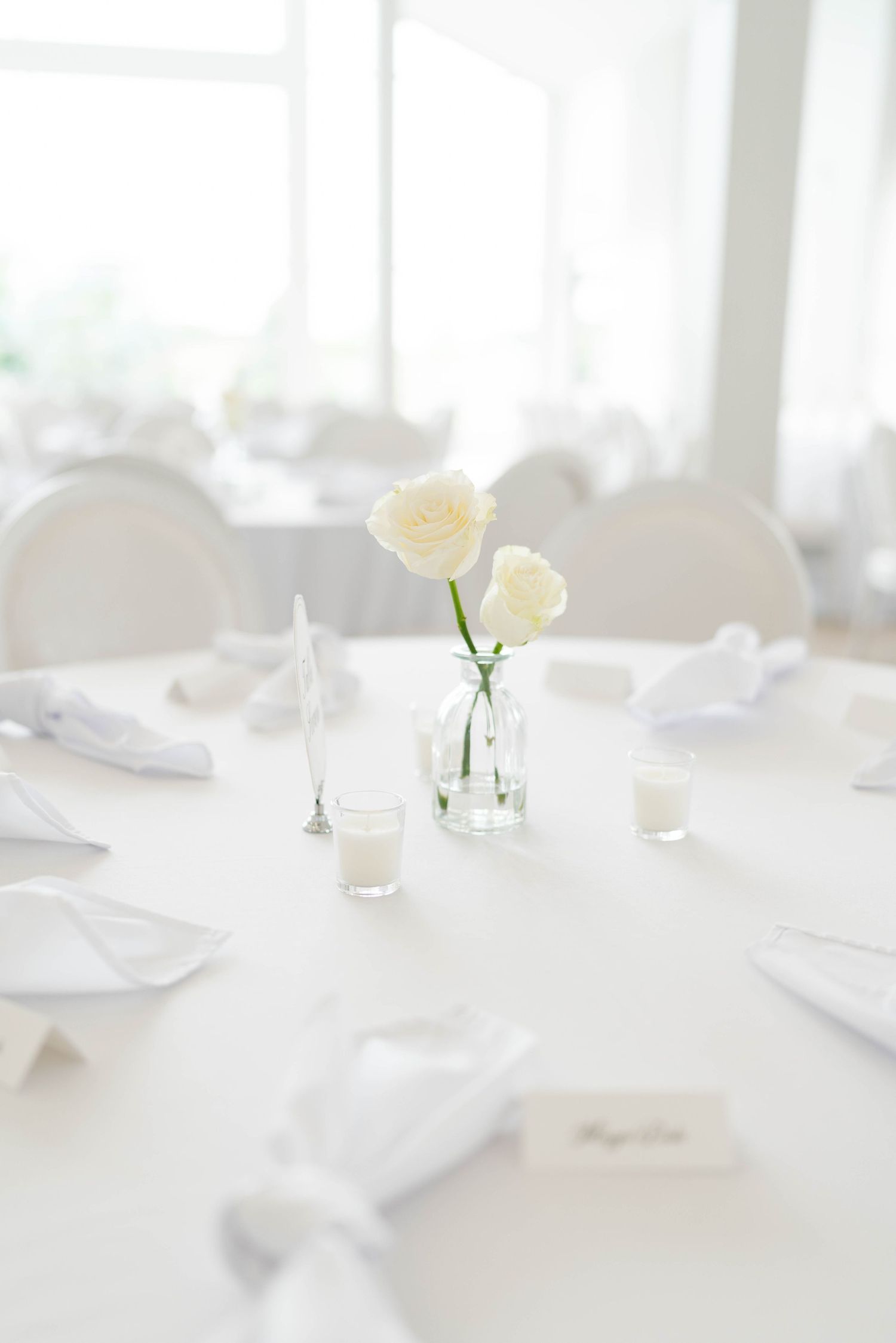 Elegant white wedding reception table with cream roses in glass bud vase, white linens, and place cards.