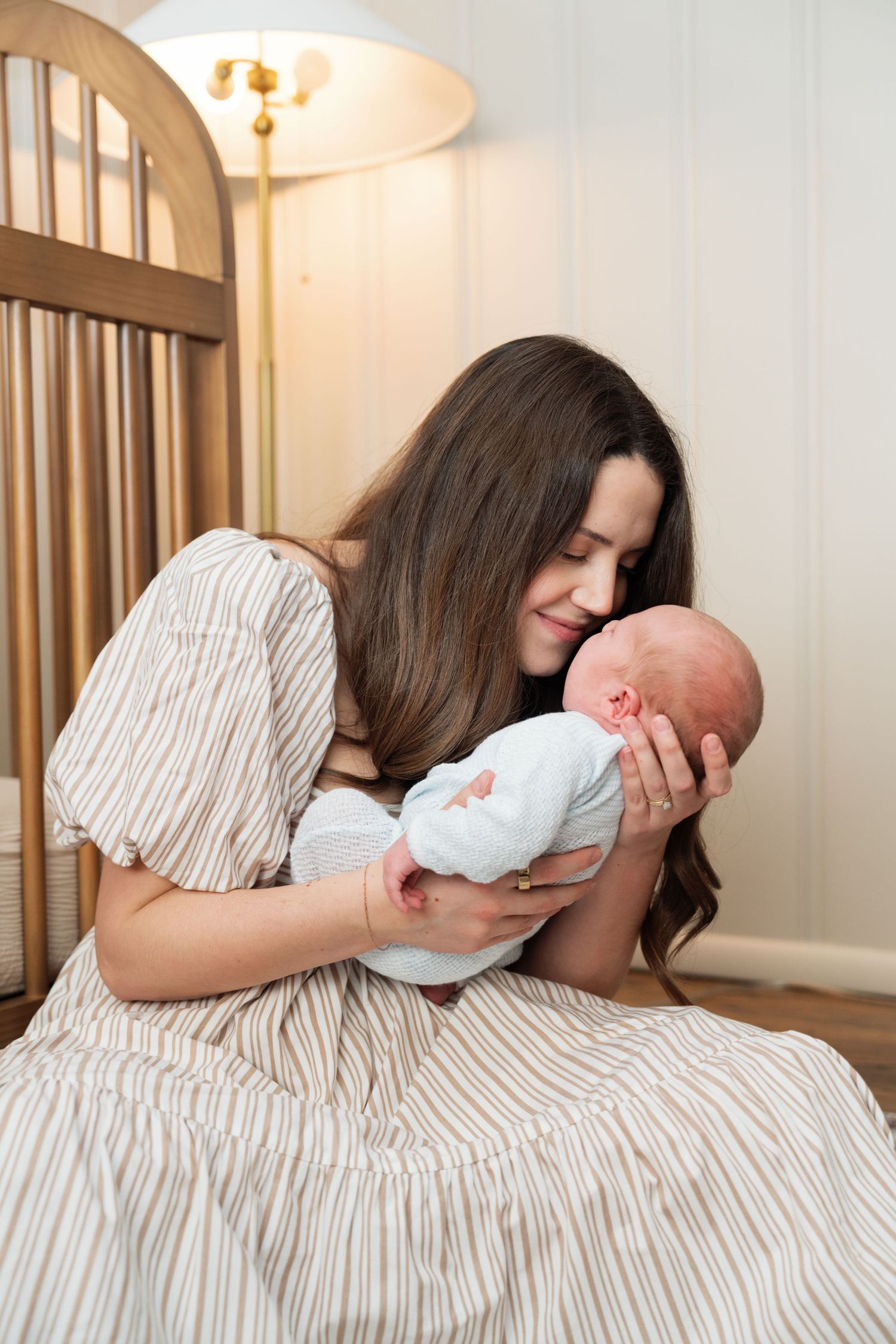 Young brunette mother smiling while cradling sleeping newborn baby in striped dress indoors.