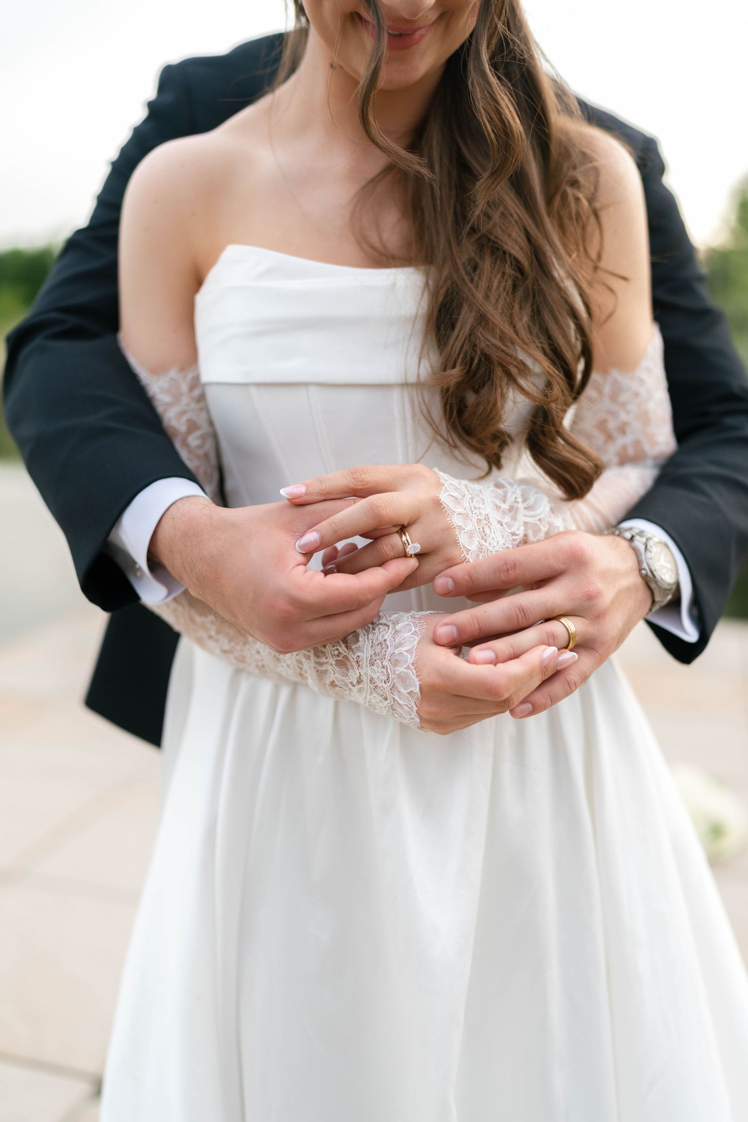 Close-up of bride and groom holding hands showing gold wedding rings, bride in strapless lace wedding gown.