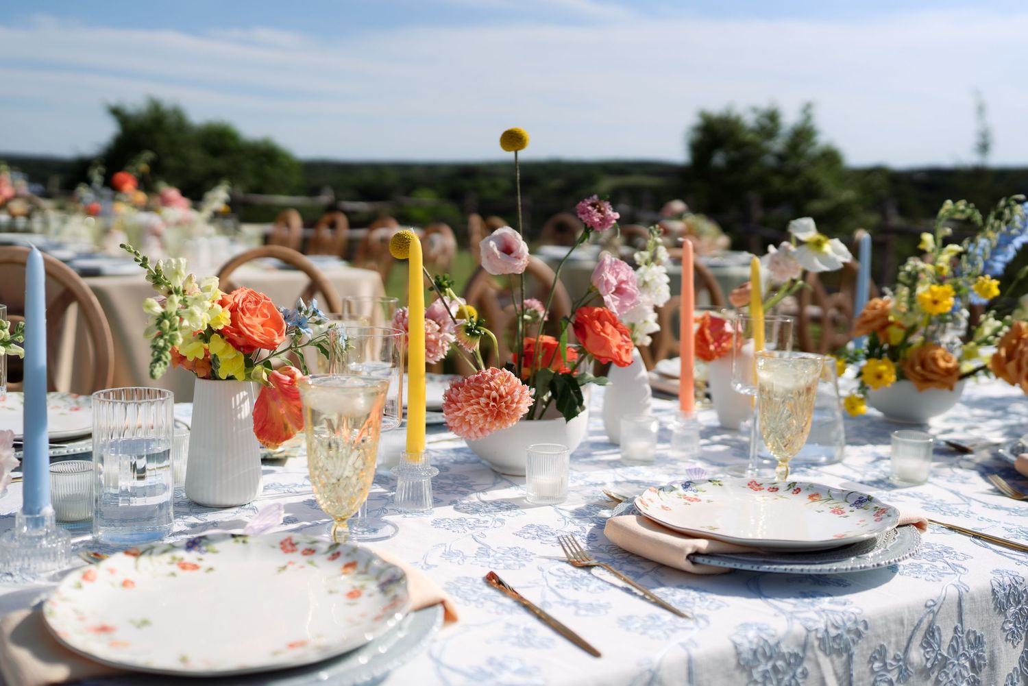 Elegant outdoor wedding tablescape with colorful floral arrangements, taper candles, and patterned dinnerware on a floral tablecloth.