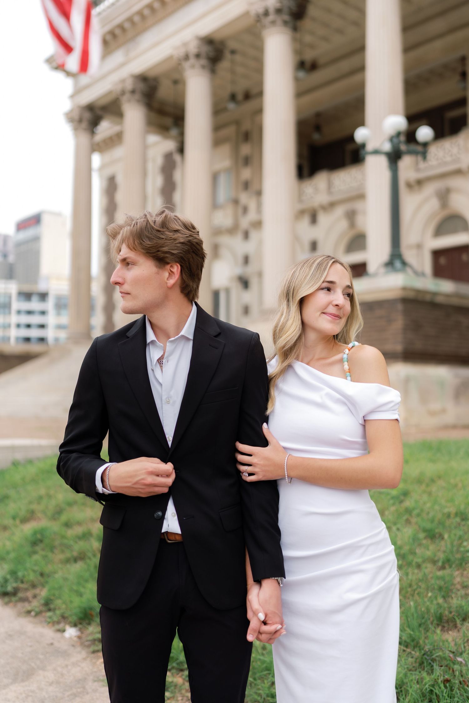 Elegant couple in formal attire holding hands outside a classical building with American flag.