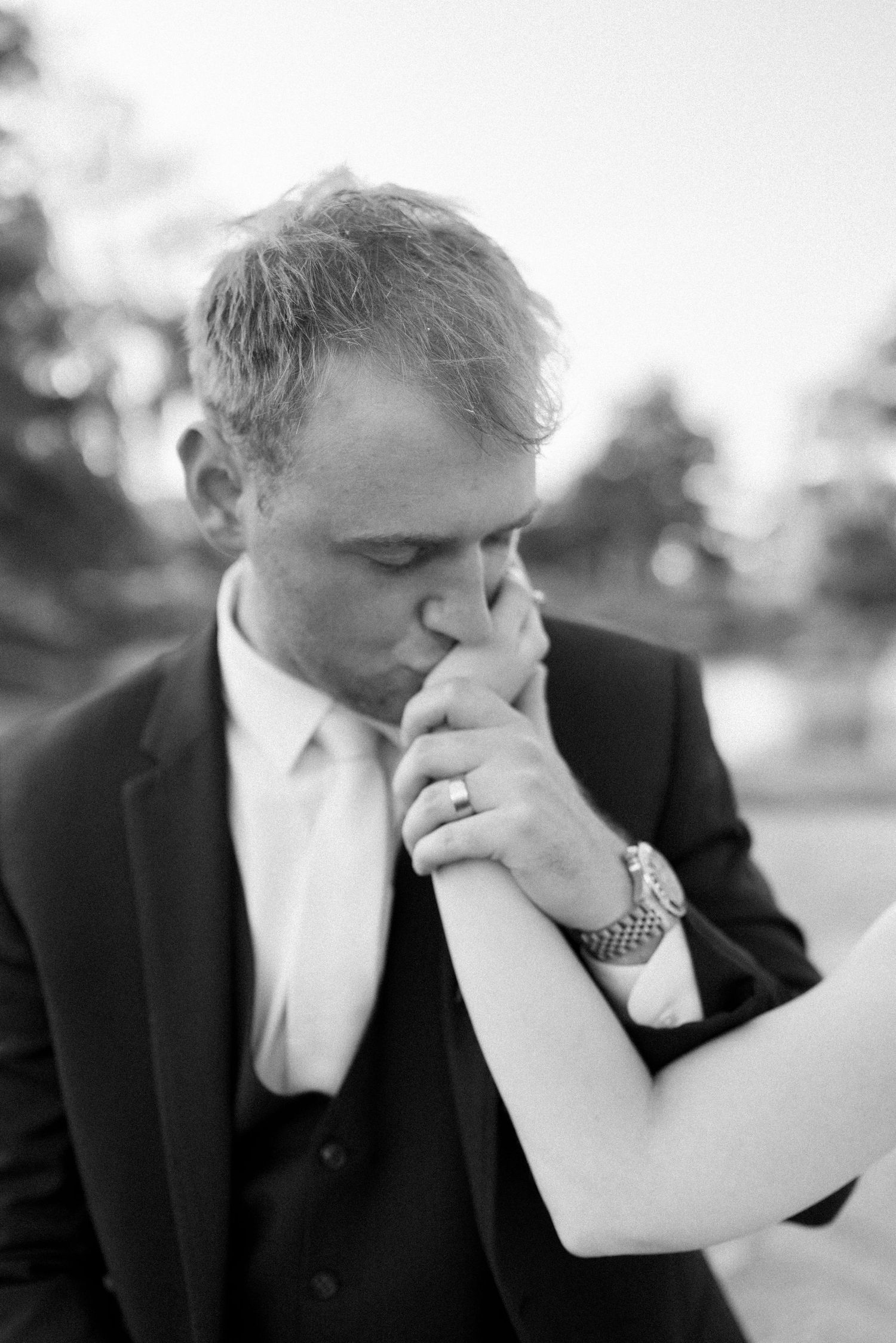 Black and white photo of groom in suit tenderly kissing bride's hand wearing a watch and wedding ring.