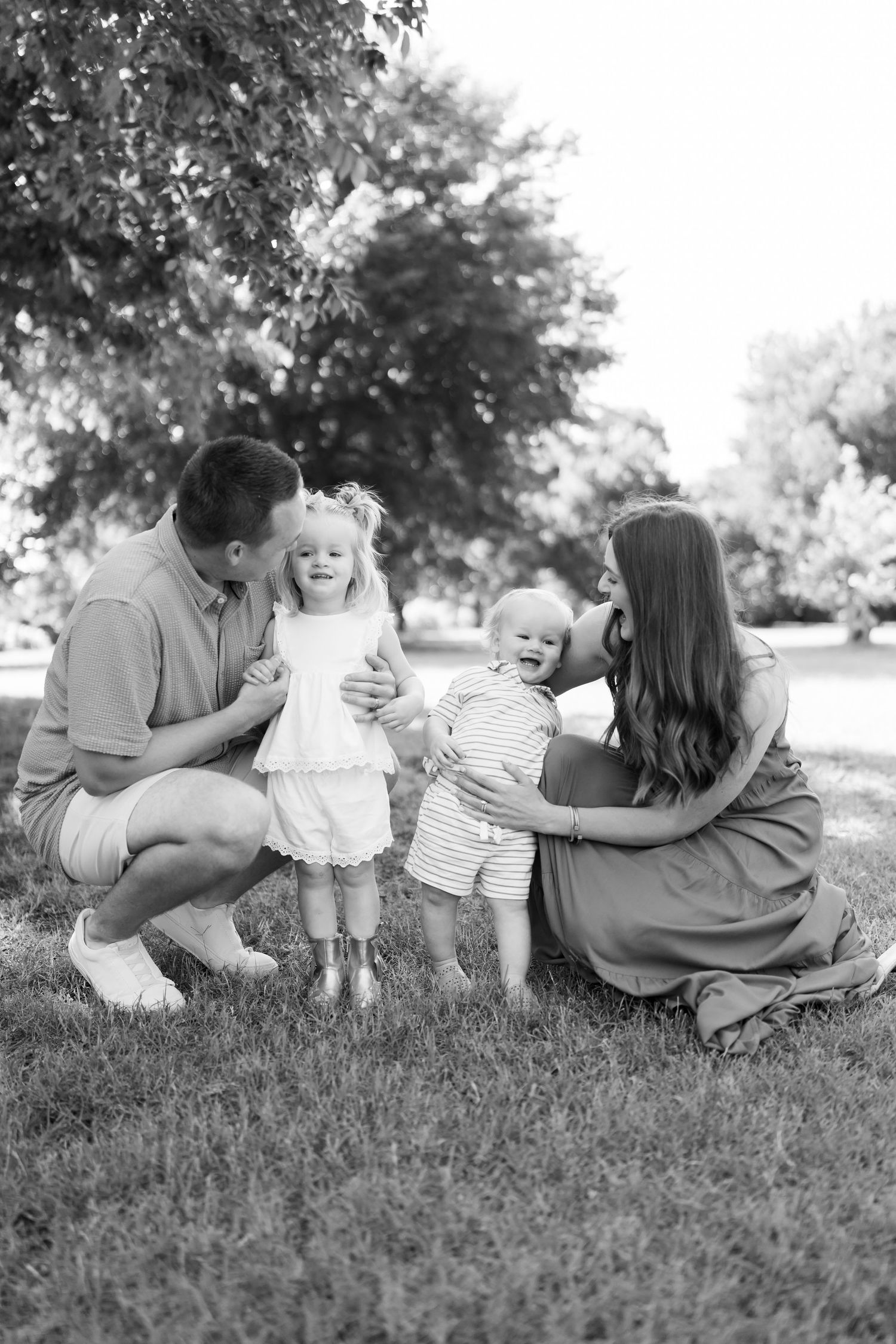 Black and white family portrait of parents with two young children laughing together on green grass outdoors.