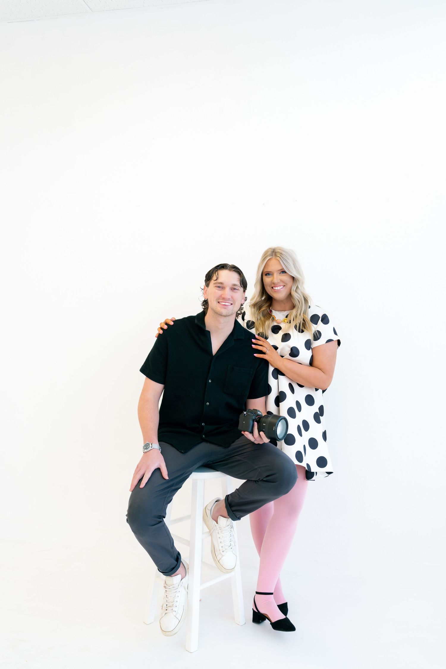 Smiling man and woman photographer duo posing together in bright white studio space.