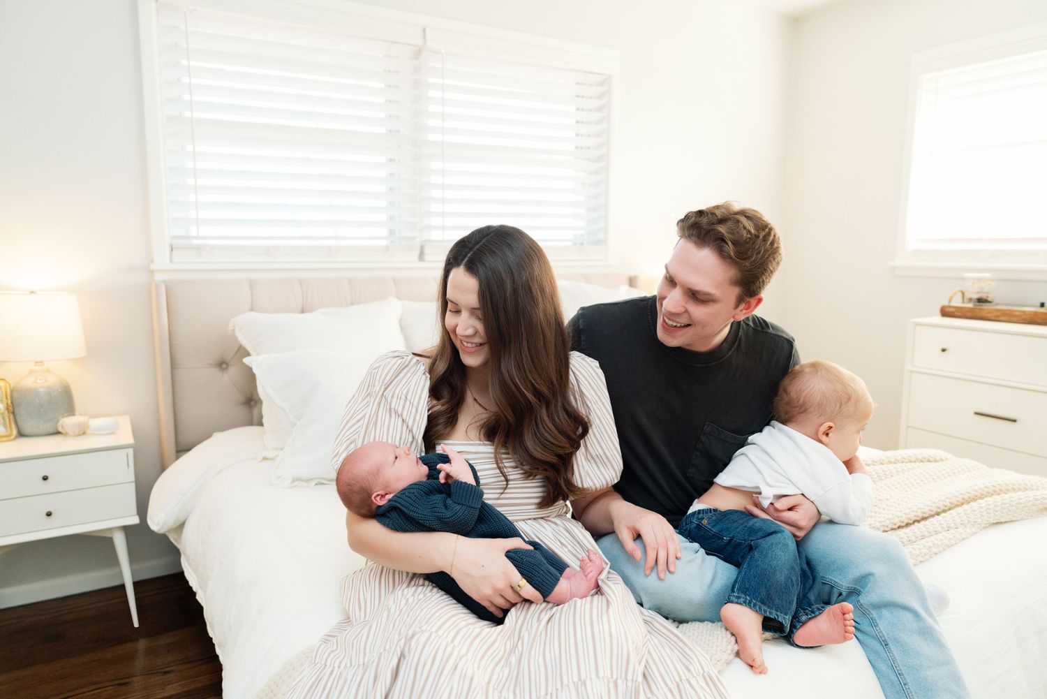 Happy family with newborn baby and toddler relaxing together on a bright white bed at home.