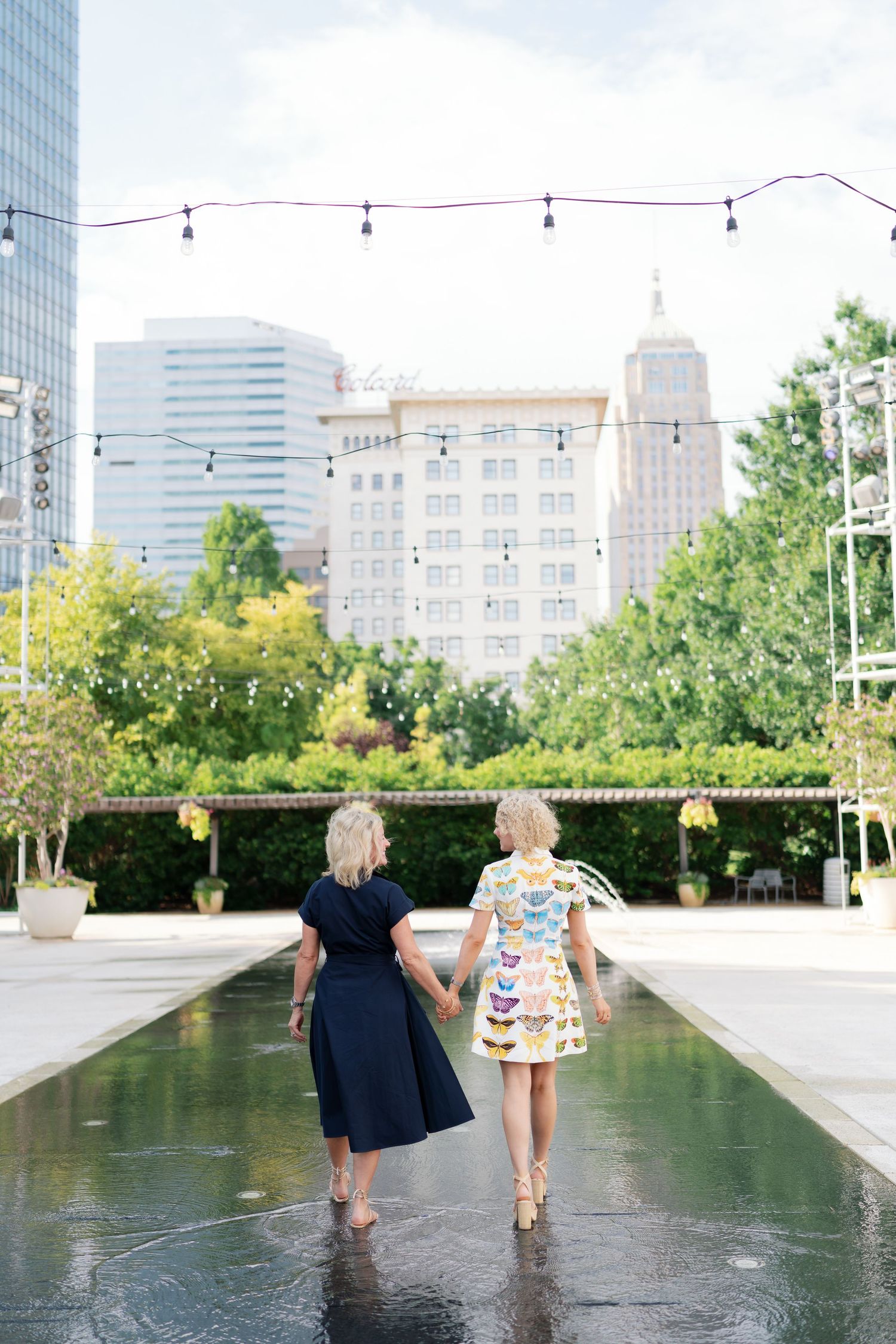 Two women in dresses holding hands walking beside a reflecting pool with city skyline and string lights above.