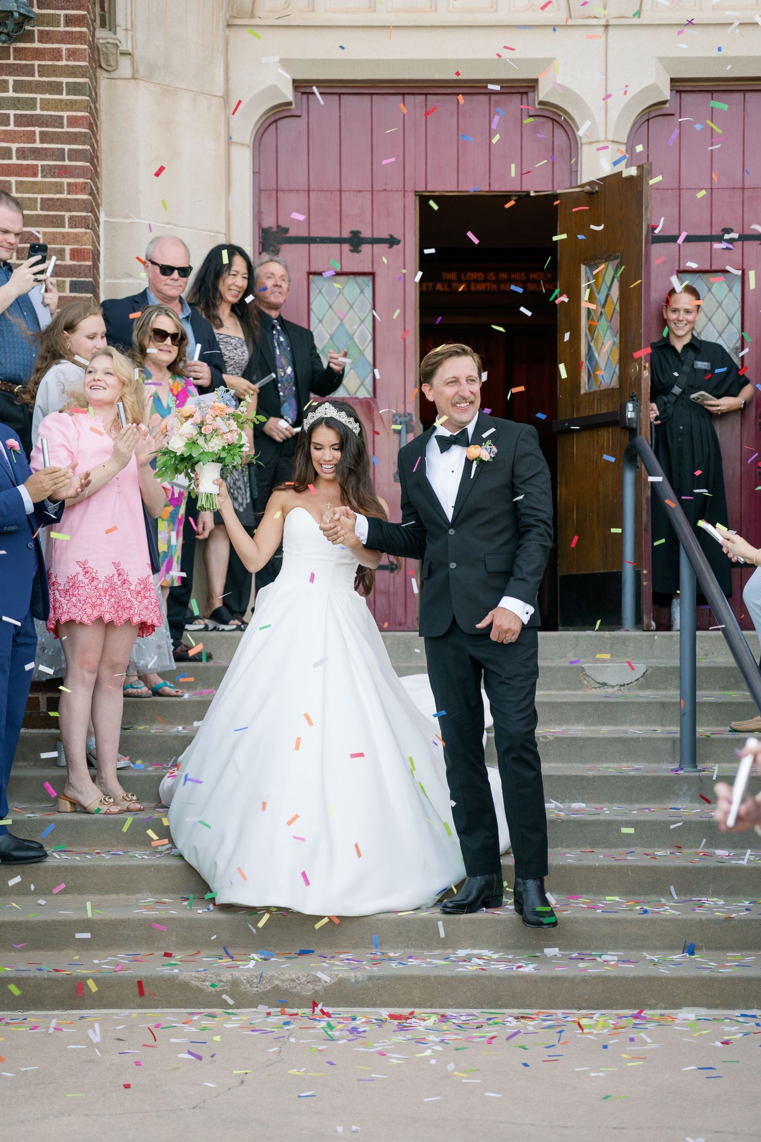 Bride and groom exit church doors as guests shower them with colorful confetti, bride in ballgown with bouquet.
