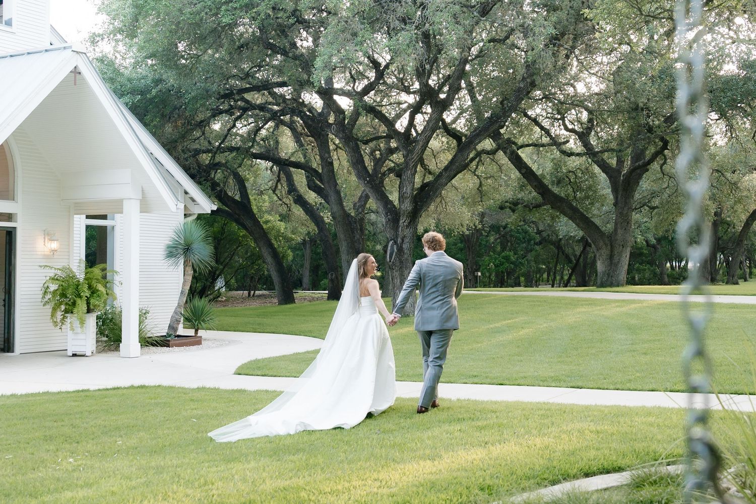 Bride and groom walking hand in hand toward a white chapel surrounded by sprawling oak trees on their wedding day.