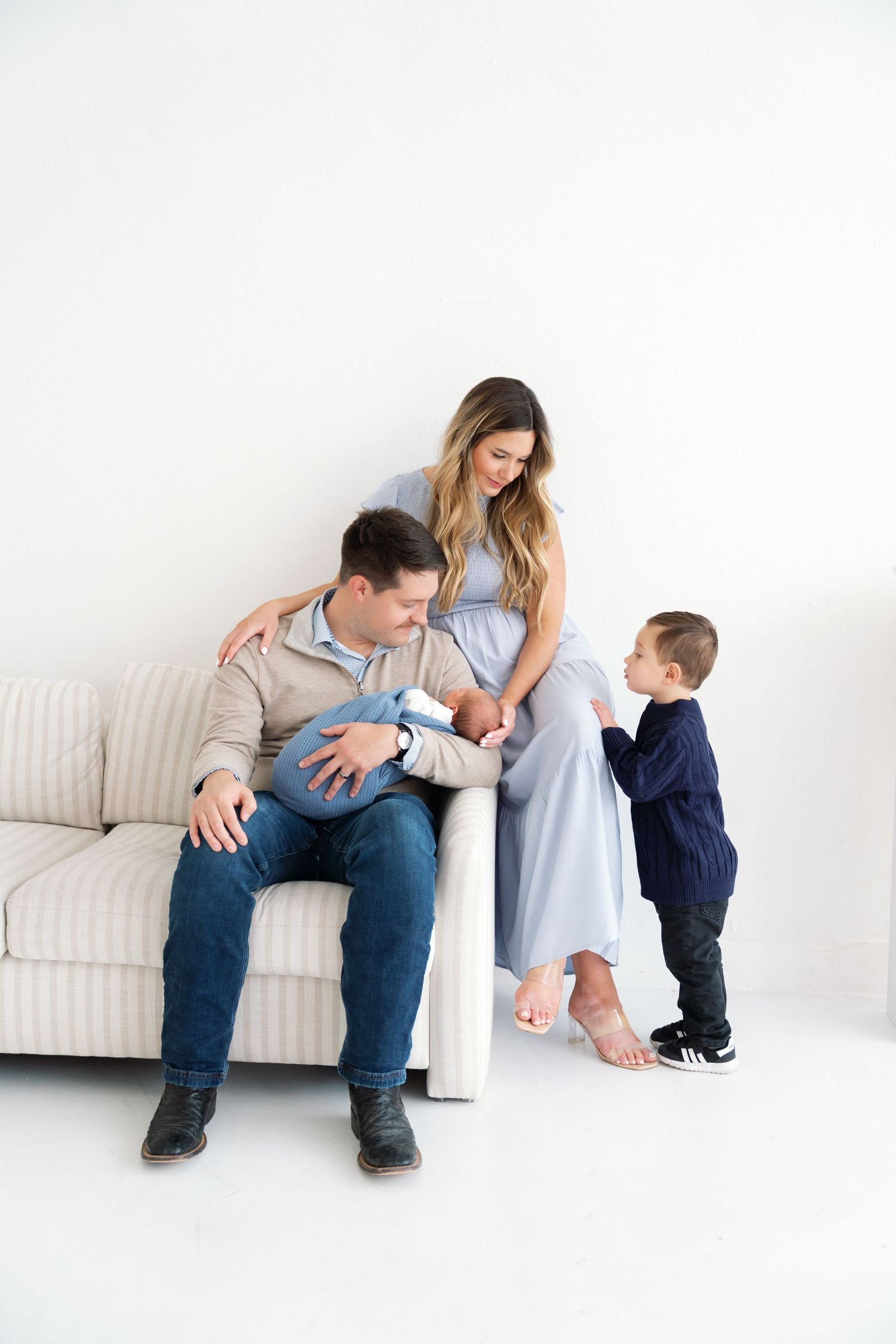 Family of four with newborn and toddler posing together on a couch in a bright white studio.