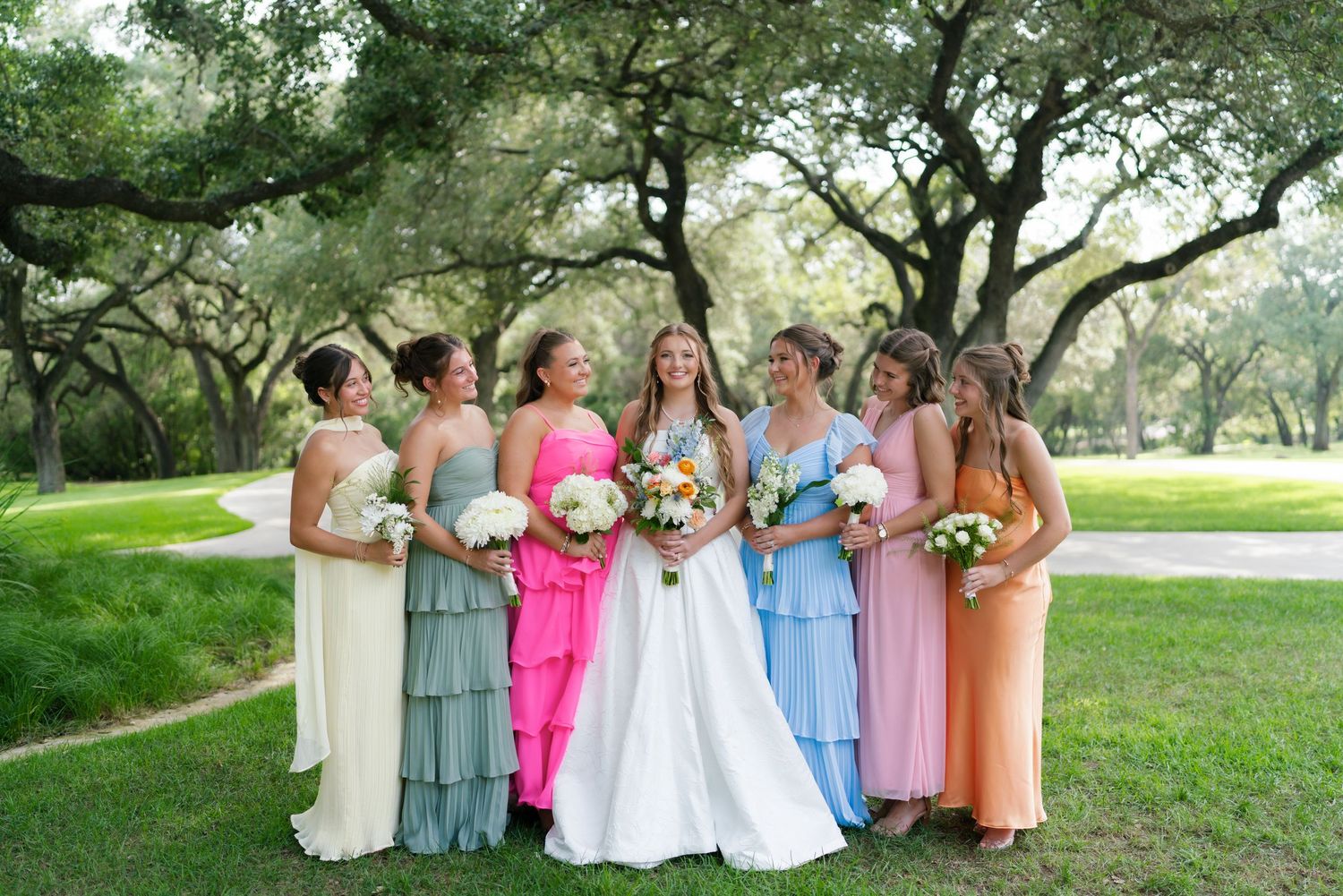 A bride poses with six bridesmaids wearing colorful mismatched pastel gowns under oak trees at an outdoor wedding.