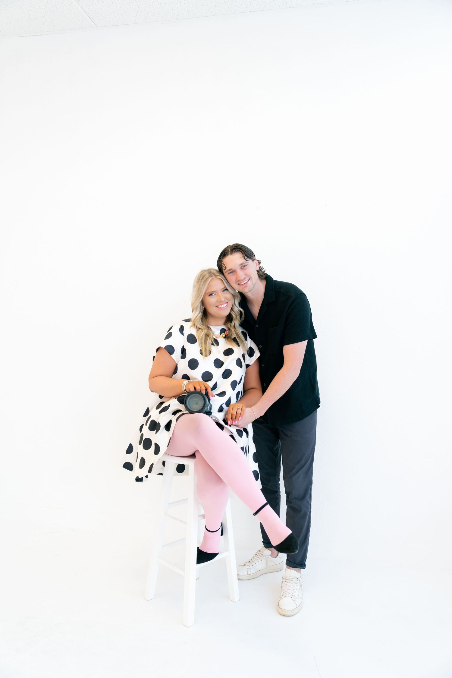 Smiling couple posing in white studio, woman in polka dot dress with pink leggings holding camera.