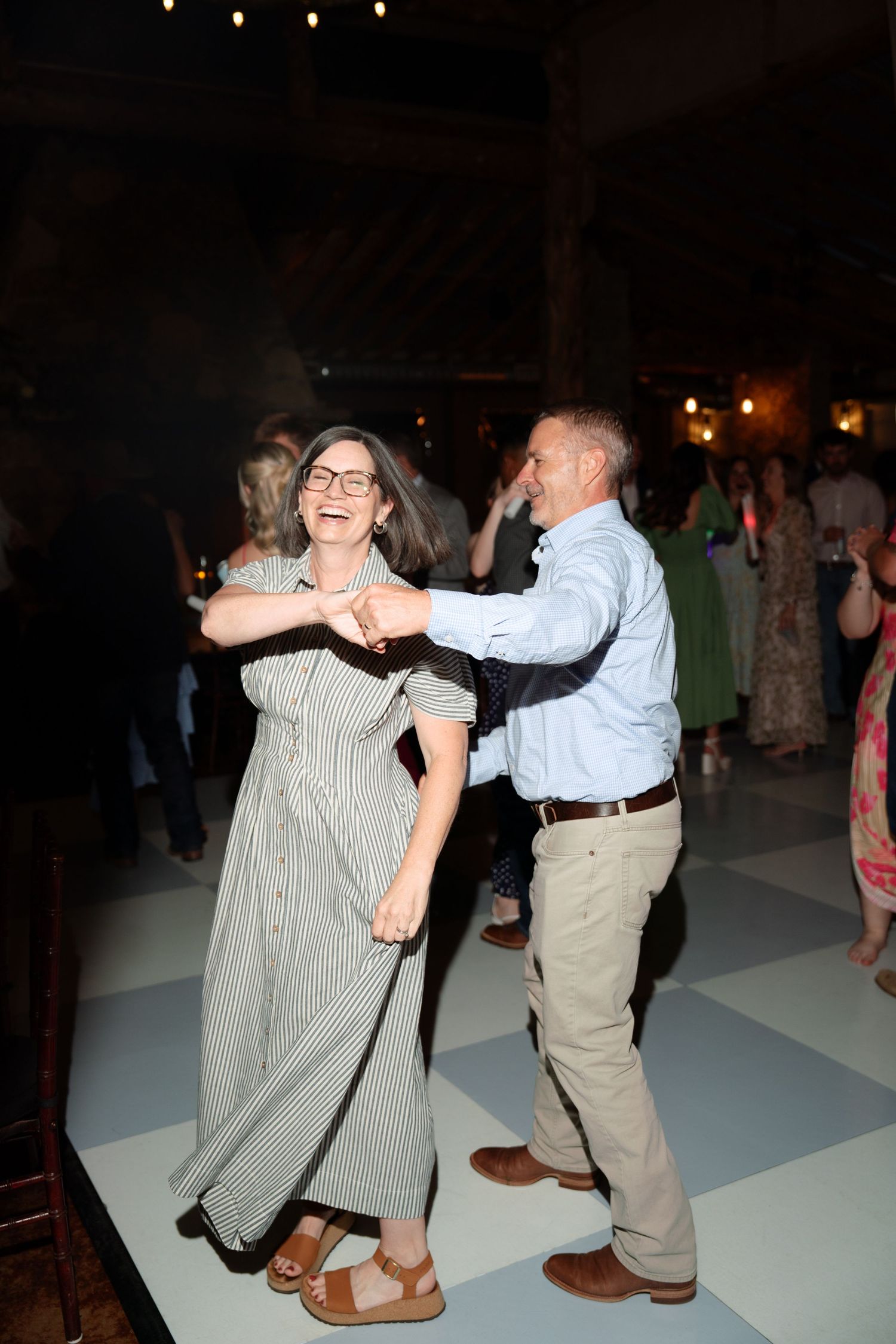 Couple dancing joyfully at an indoor event, woman in striped dress laughing, man in blue shirt on dance floor.
