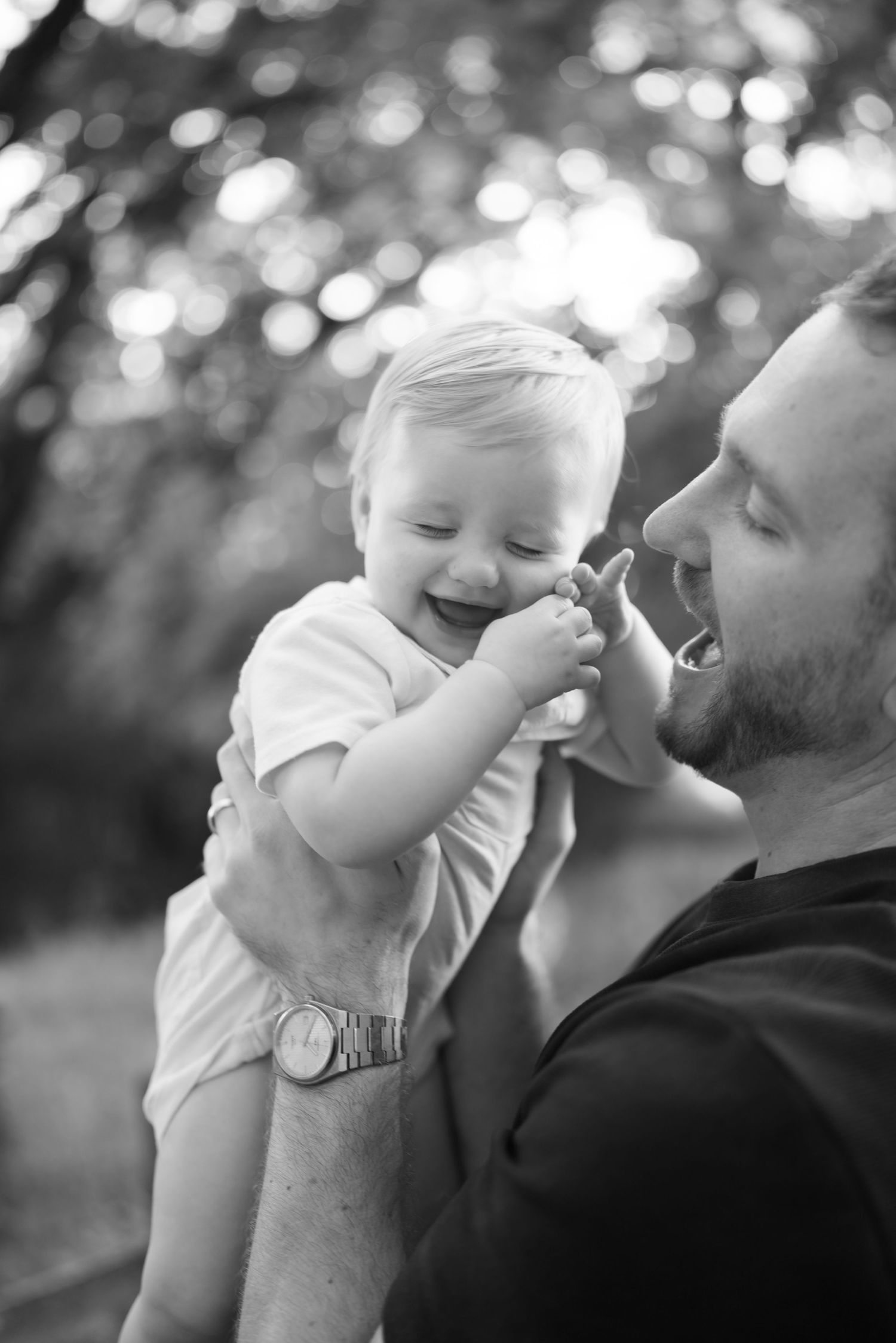 Black and white photo of a joyful baby laughing while being held by a bearded father outdoors.