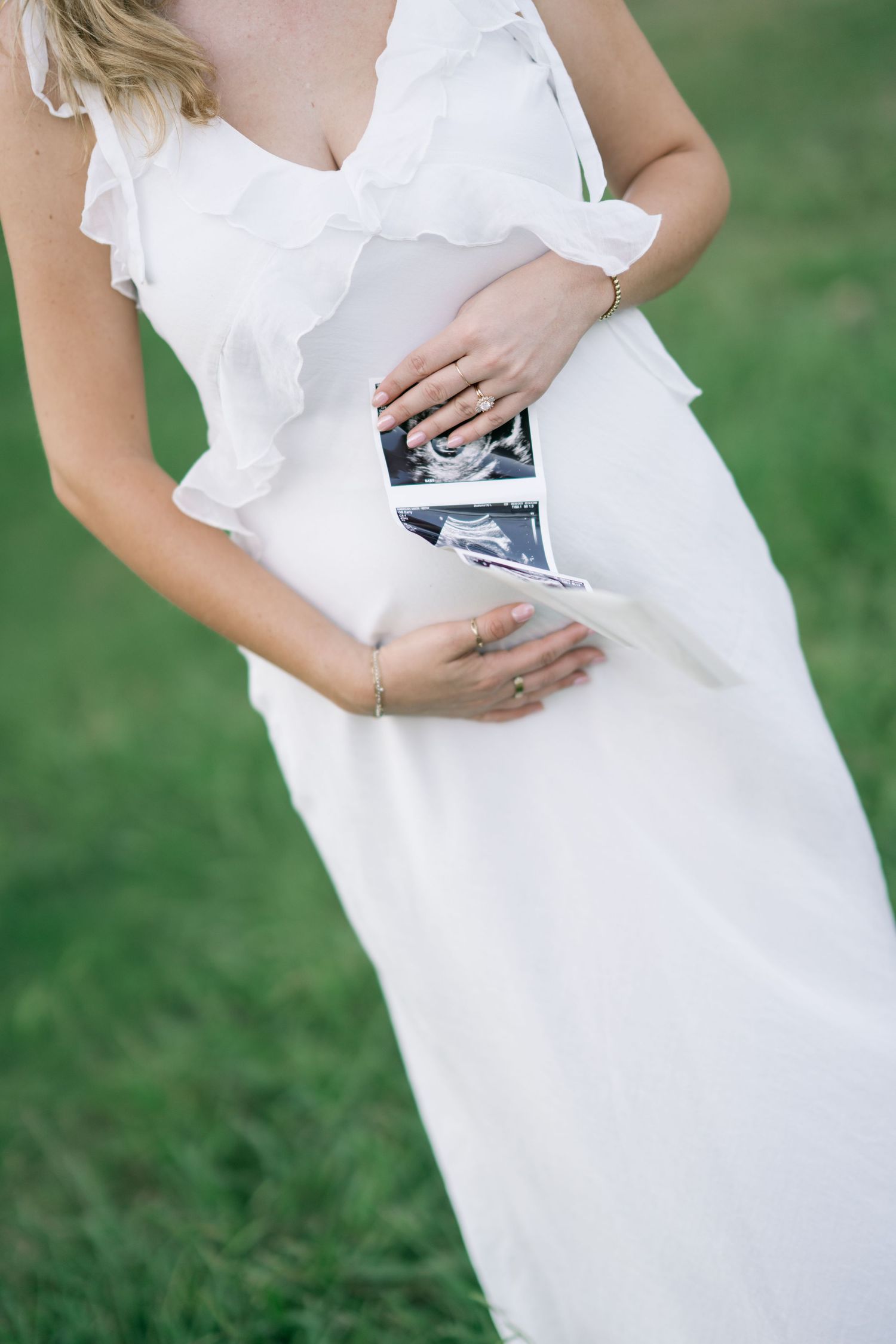 Pregnant woman in white dress holding ultrasound photos against her baby bump outdoors.