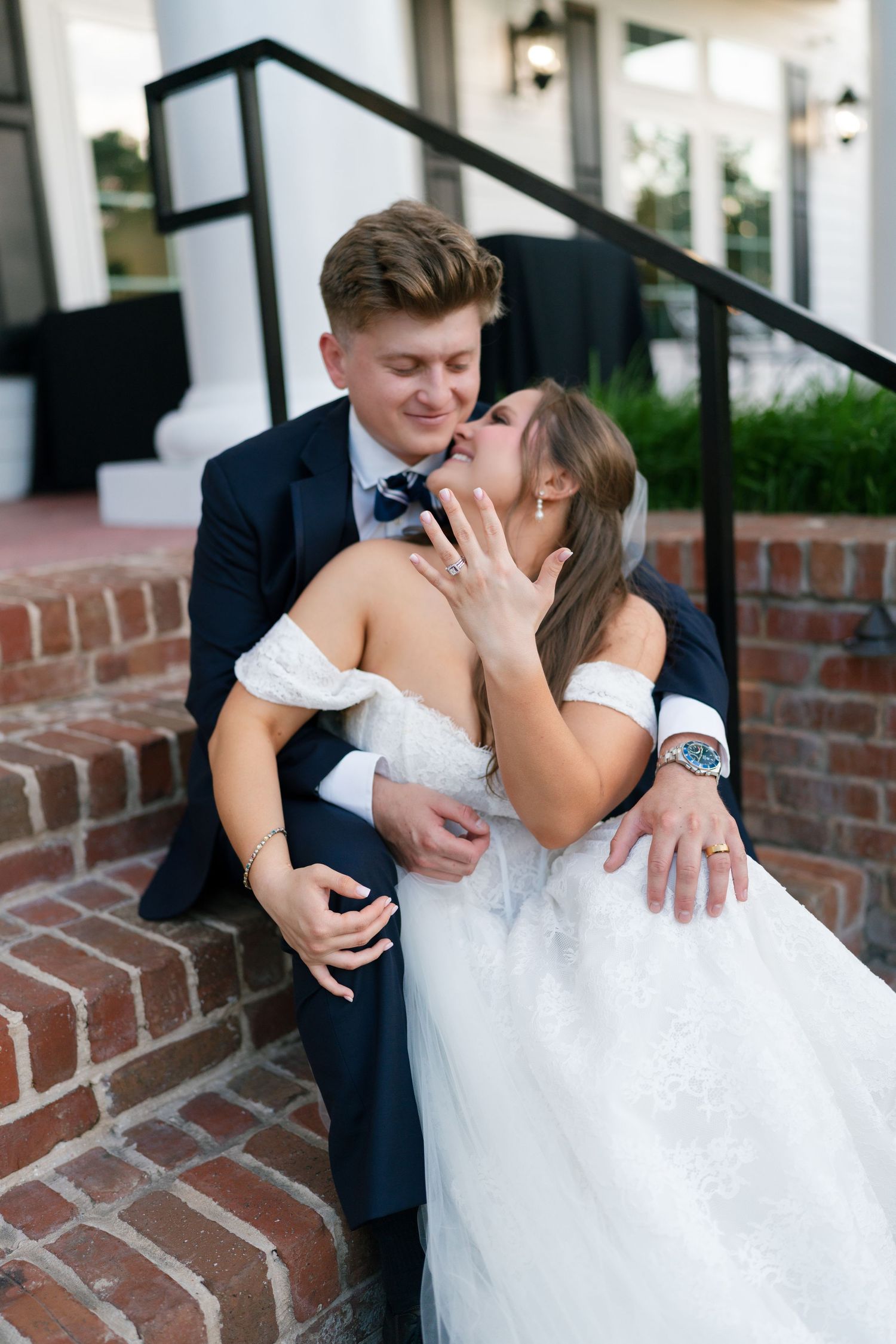 Bride and groom sharing a kiss on brick steps, bride showing off wedding ring in white off-shoulder gown and navy suit.