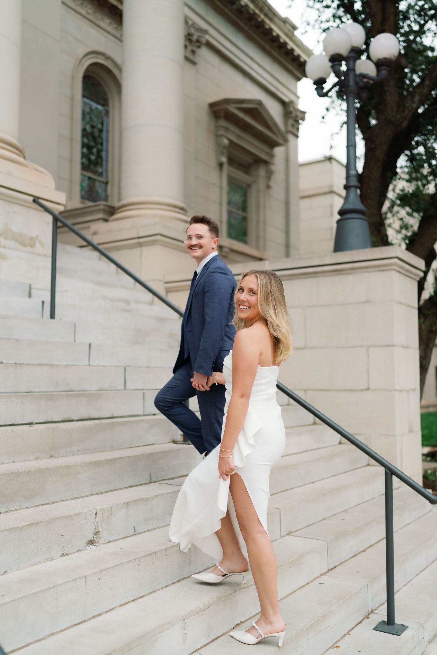 Bride and groom smiling on stone steps of a grand building, bride in white dress and groom in navy suit.