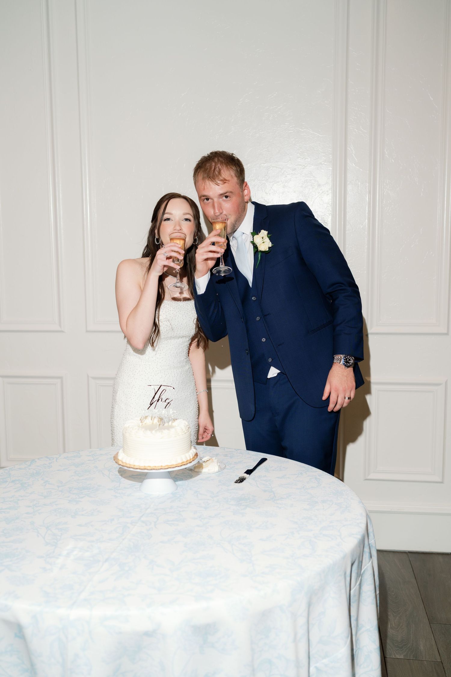 Bride and groom toasting champagne beside white two-tier wedding cake, groom in navy suit with floral boutonniere.