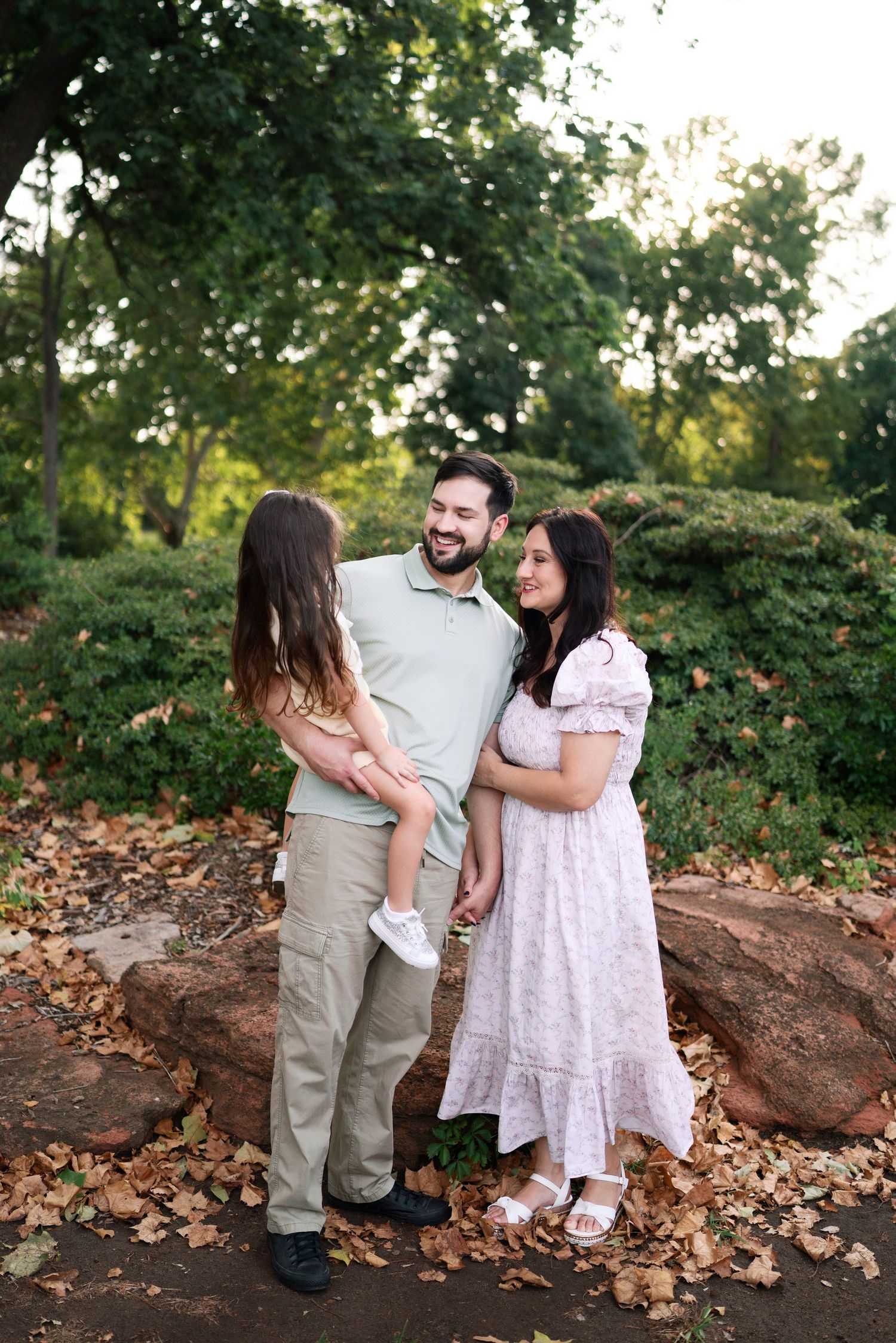 Smiling family trio standing together on rocks covered in autumn leaves in a green park setting.