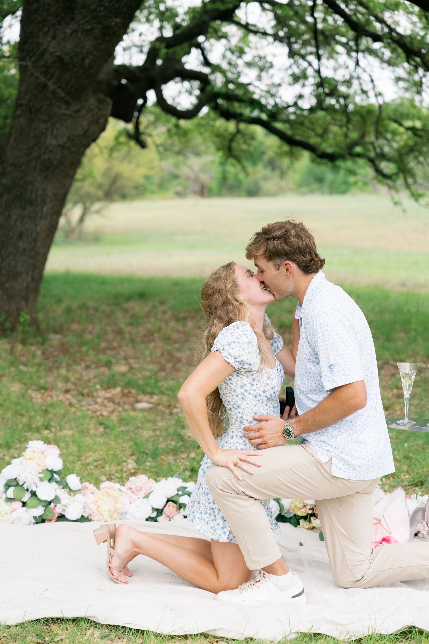 Couple sharing a kiss during a romantic outdoor picnic proposal surrounded by flowers and champagne.