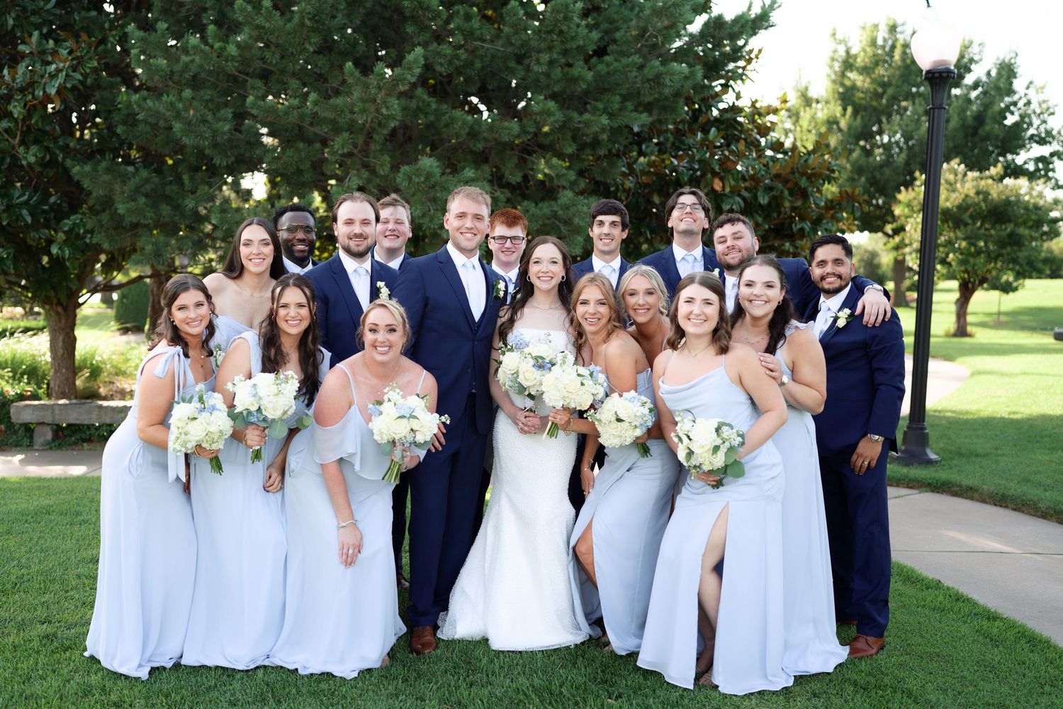 Large wedding party poses on green lawn, bridesmaids in blue gowns and groomsmen in navy suits.