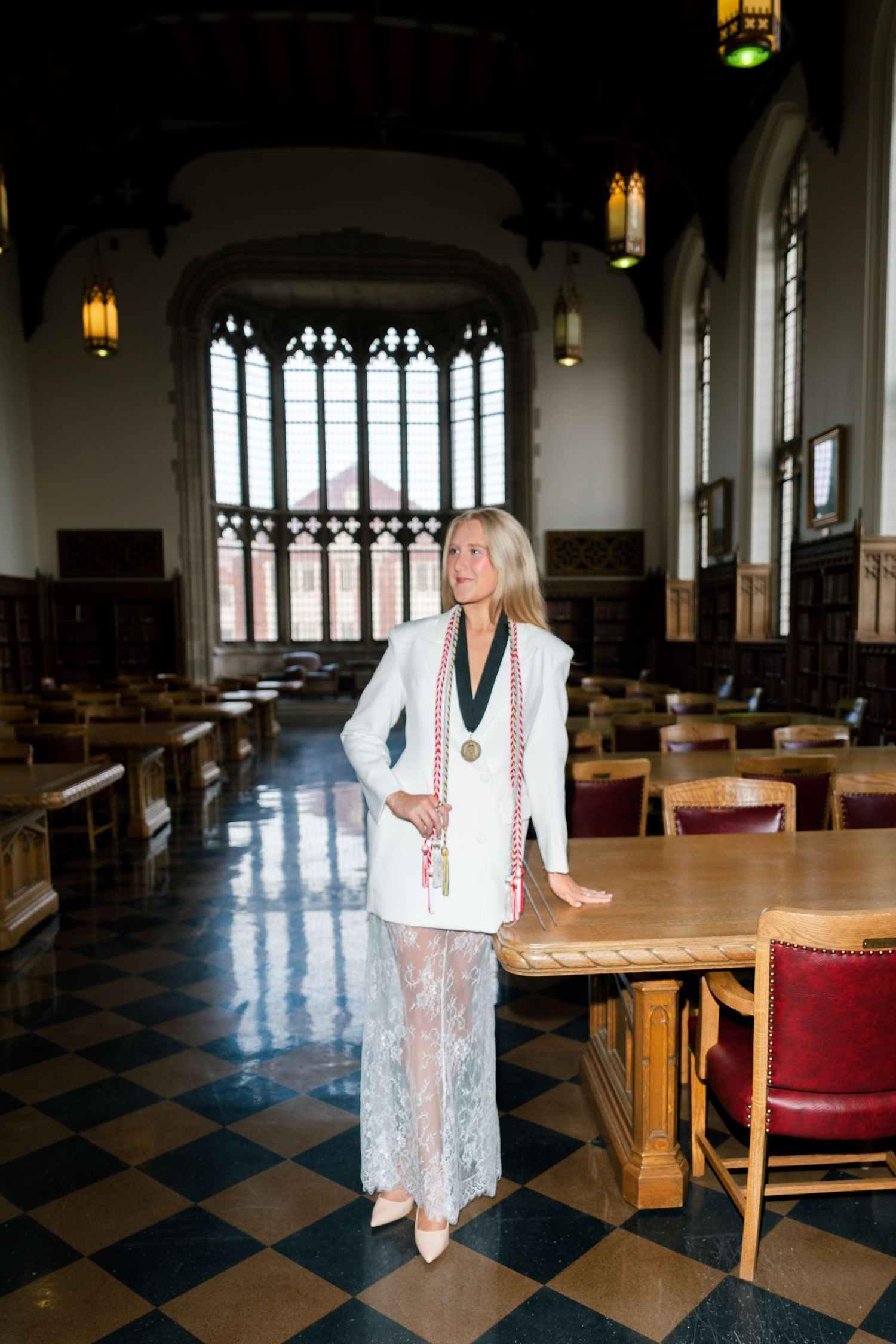 Blonde woman in white blazer and lace pants posing in elegant university hall with stained glass windows.
