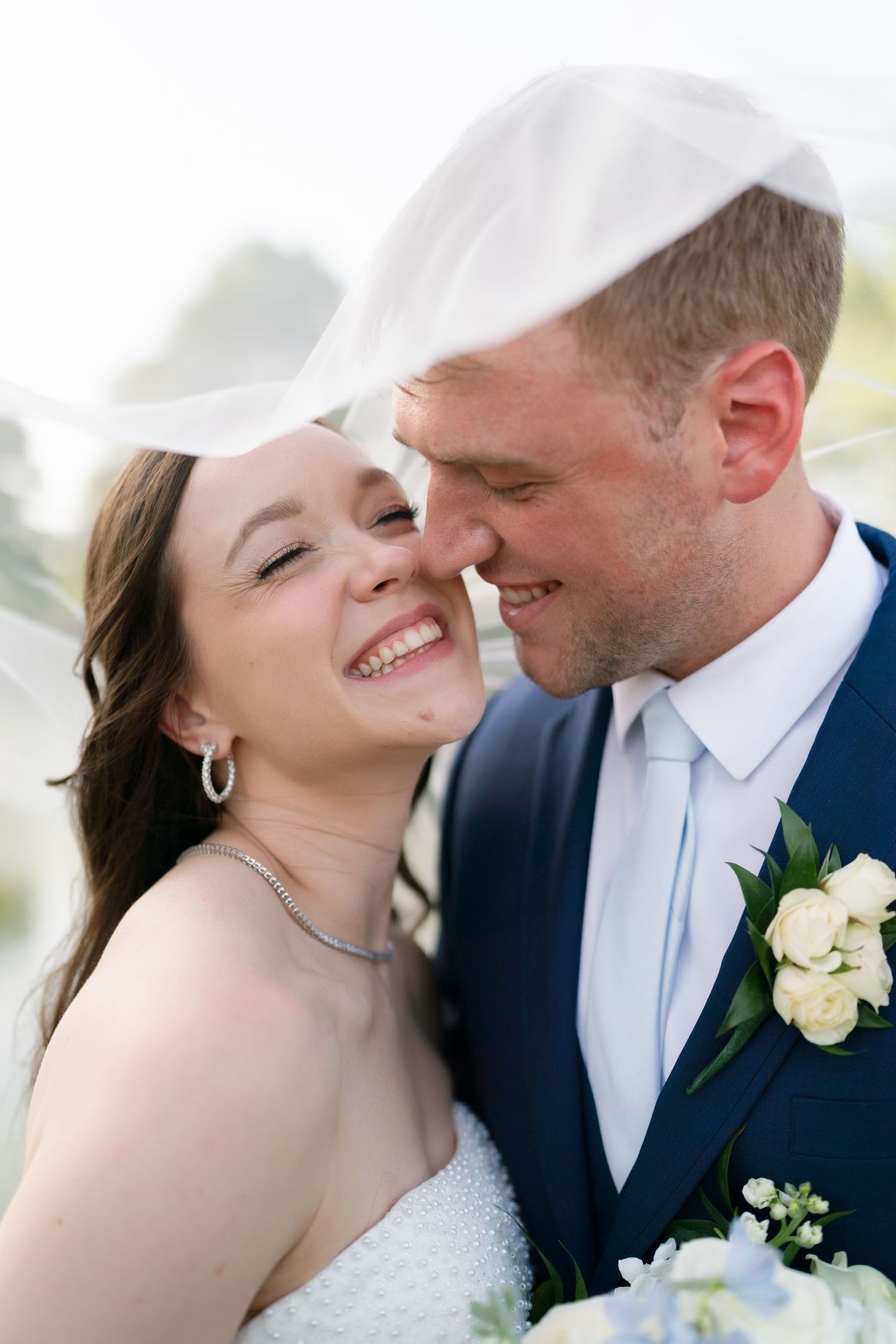 Bride and groom sharing a romantic moment under a flowing white veil, groom in navy suit with bouquet.