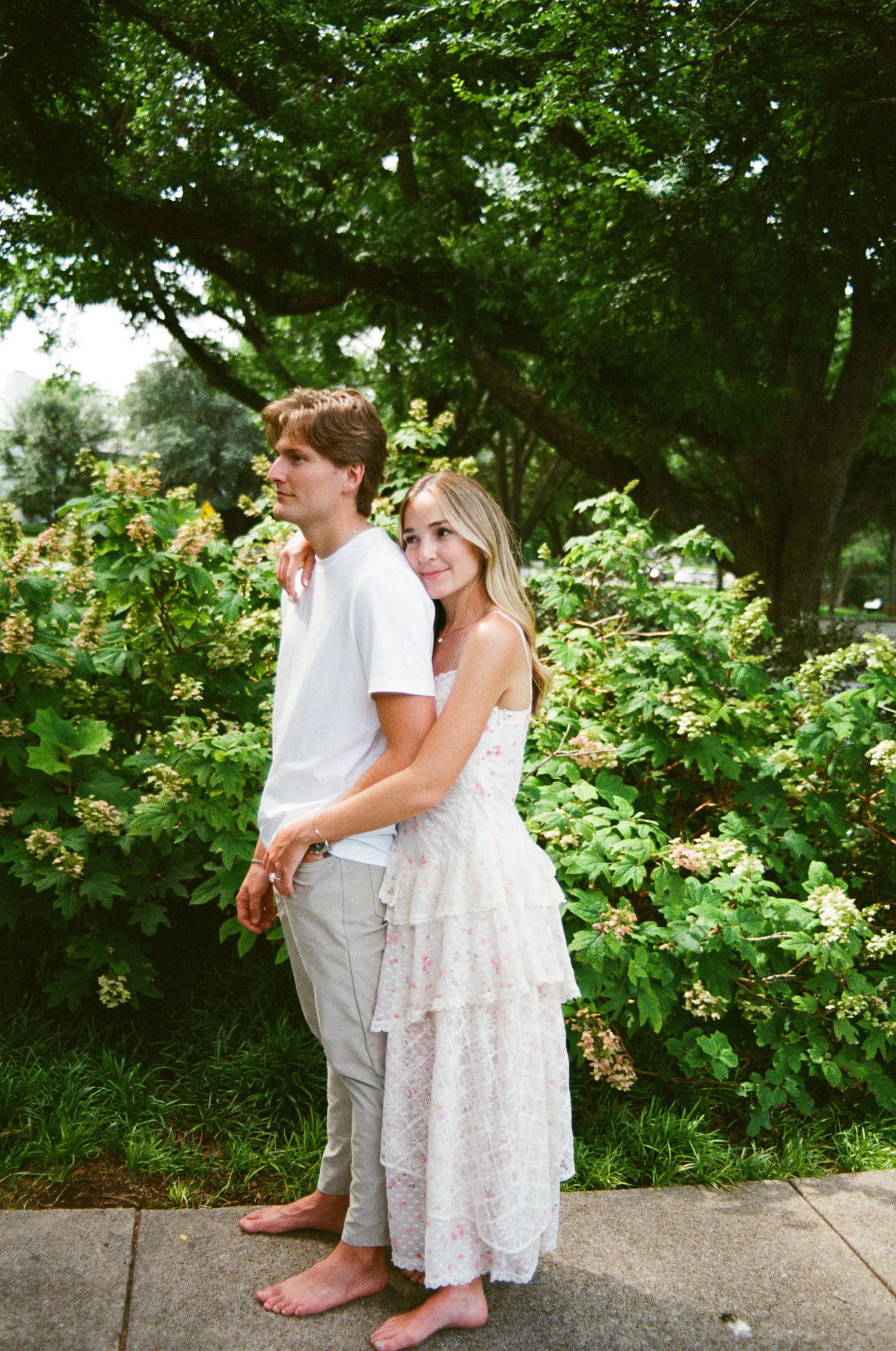 Barefoot couple poses together in front of blooming green bushes, woman in floral dress embracing man.