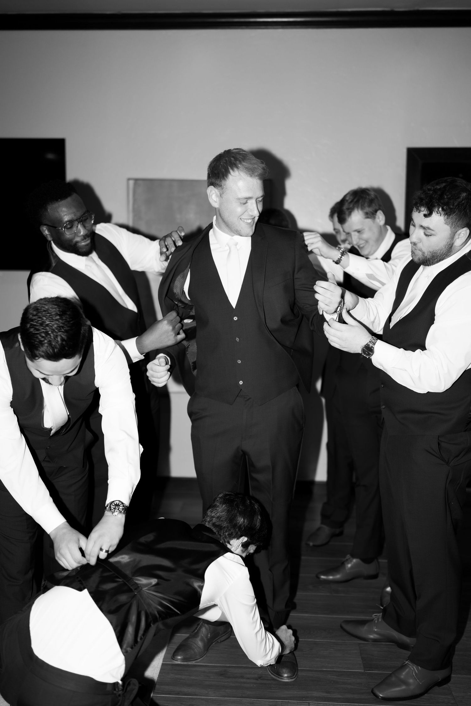 Black and white photo of a groom surrounded by groomsmen in vests helping him get dressed before the wedding.