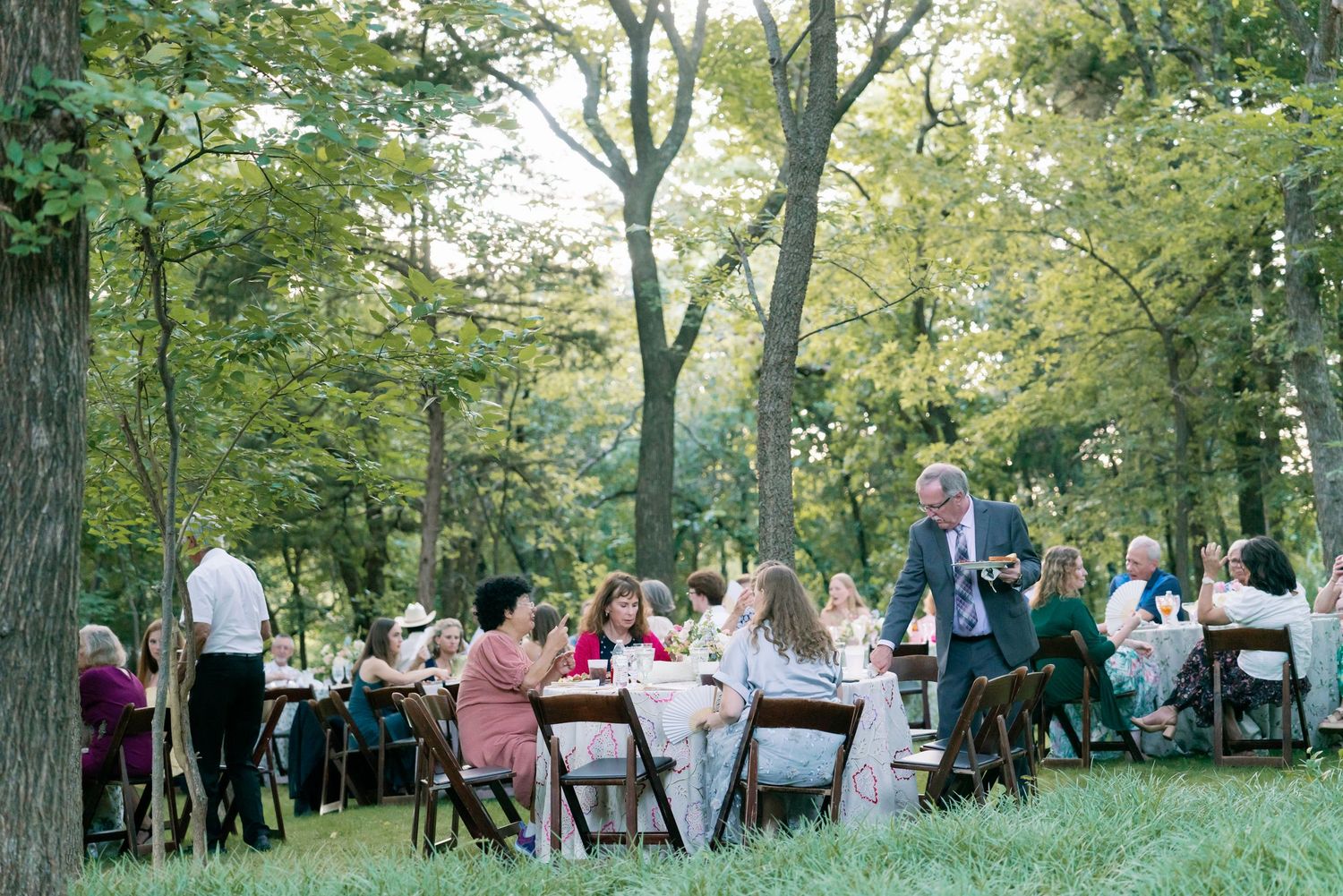 Elegant outdoor wedding reception dinner under tall trees with guests seated at long decorated tables.