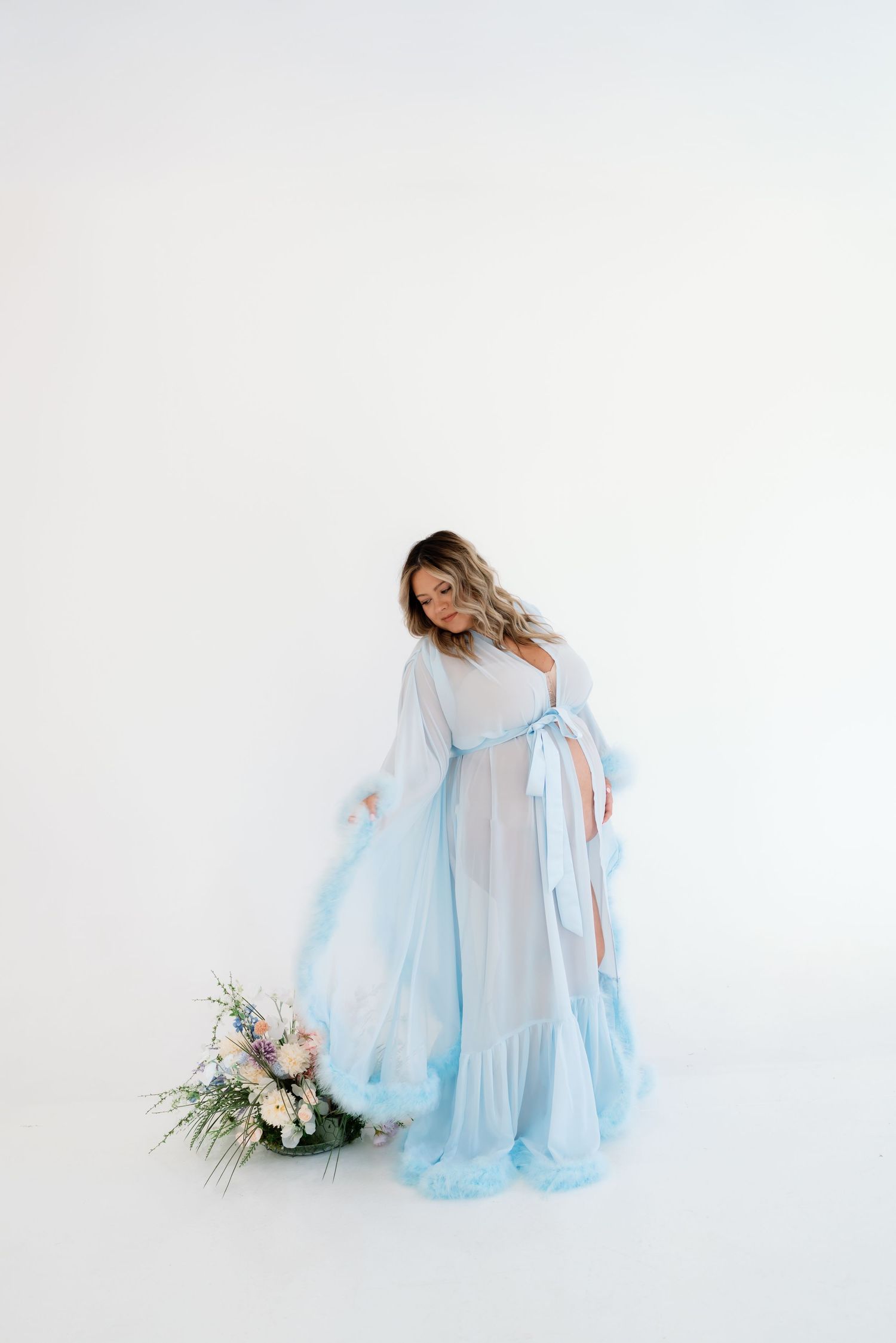 Pregnant woman in a flowing light blue sheer maternity gown standing beside a floral bouquet on white background.