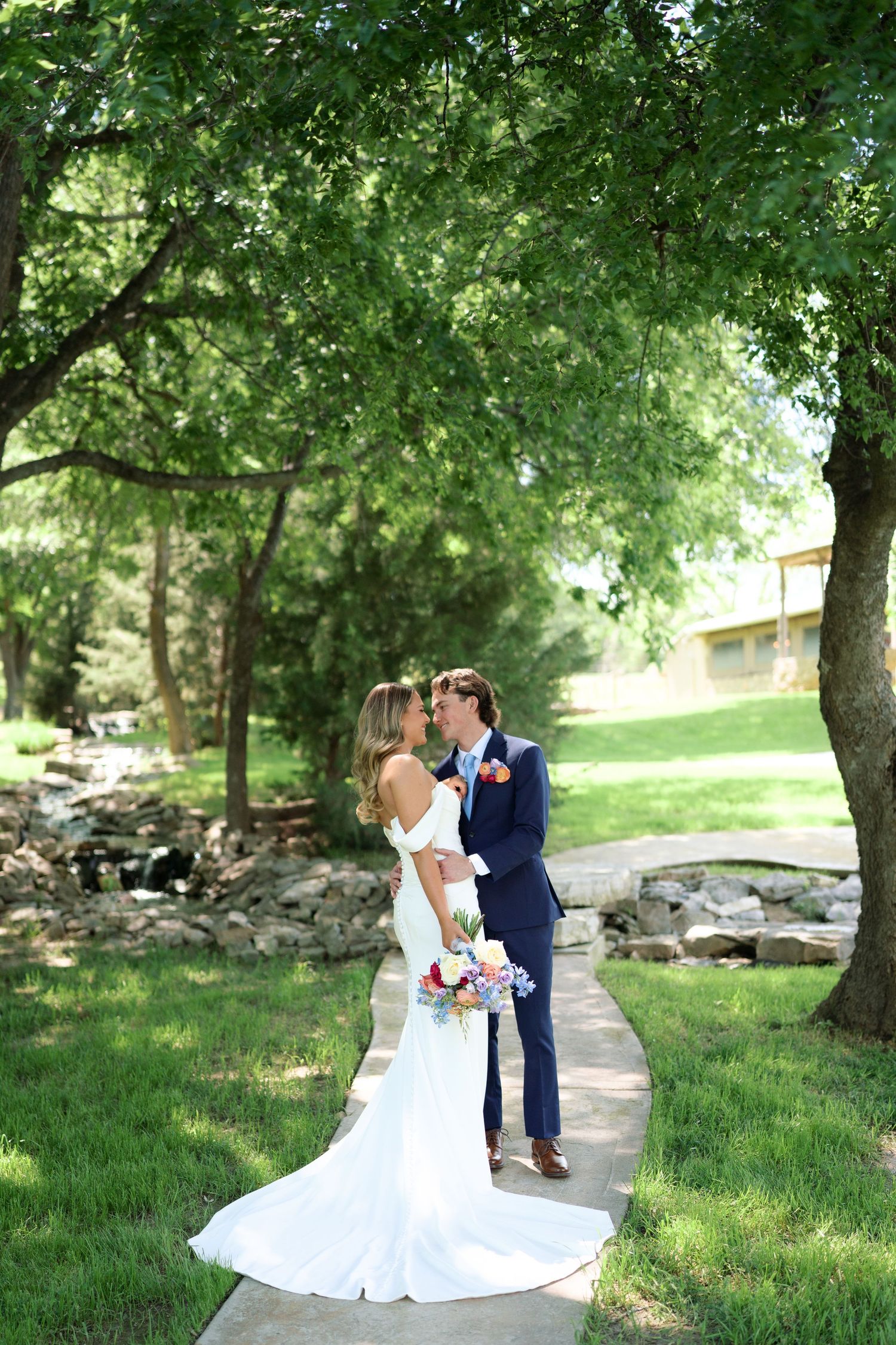 A bride and groom share a romantic kiss on a garden pathway surrounded by lush green trees at their outdoor wedding.