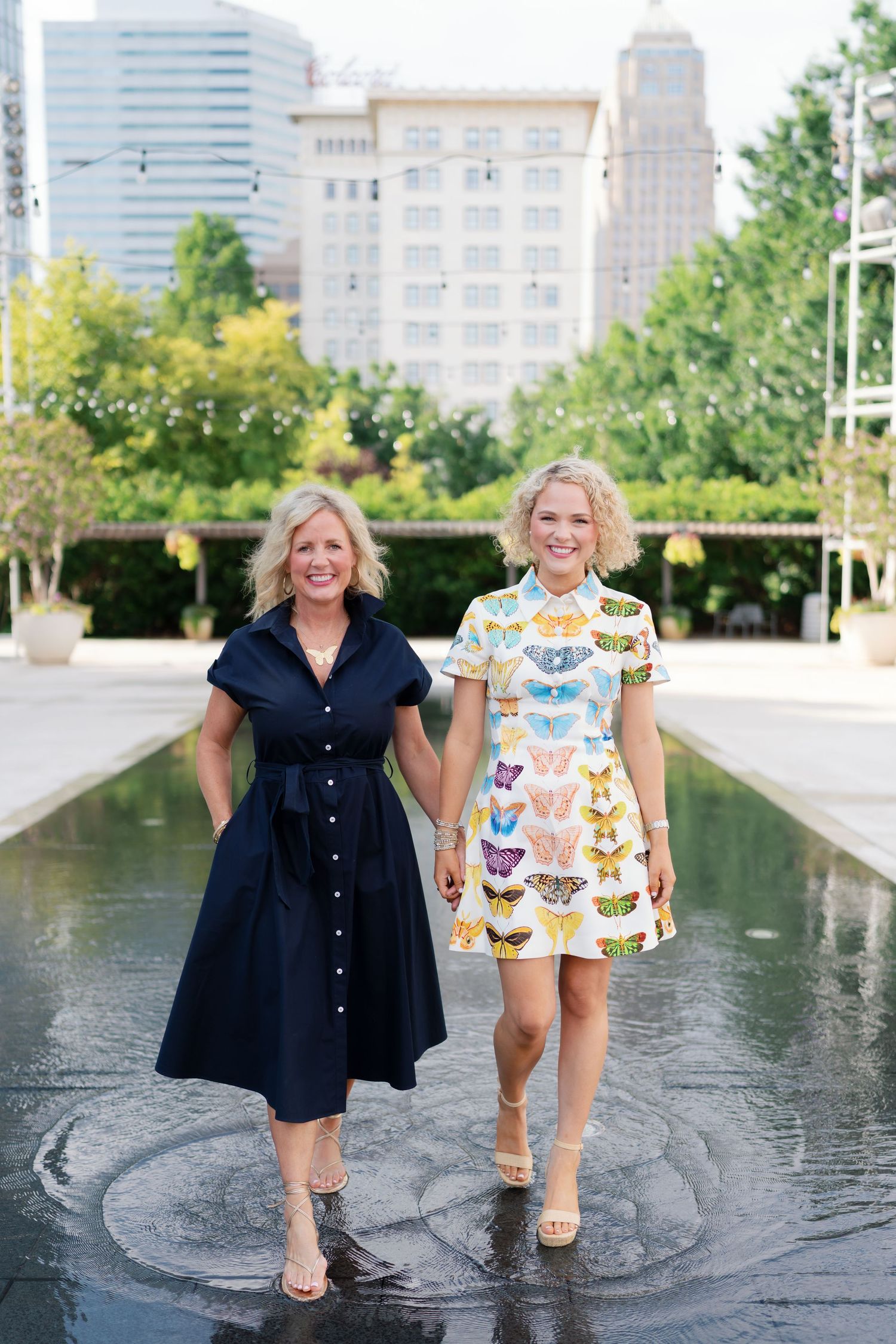 Two women in dresses hold hands walking by a reflecting pool with city buildings in the background.