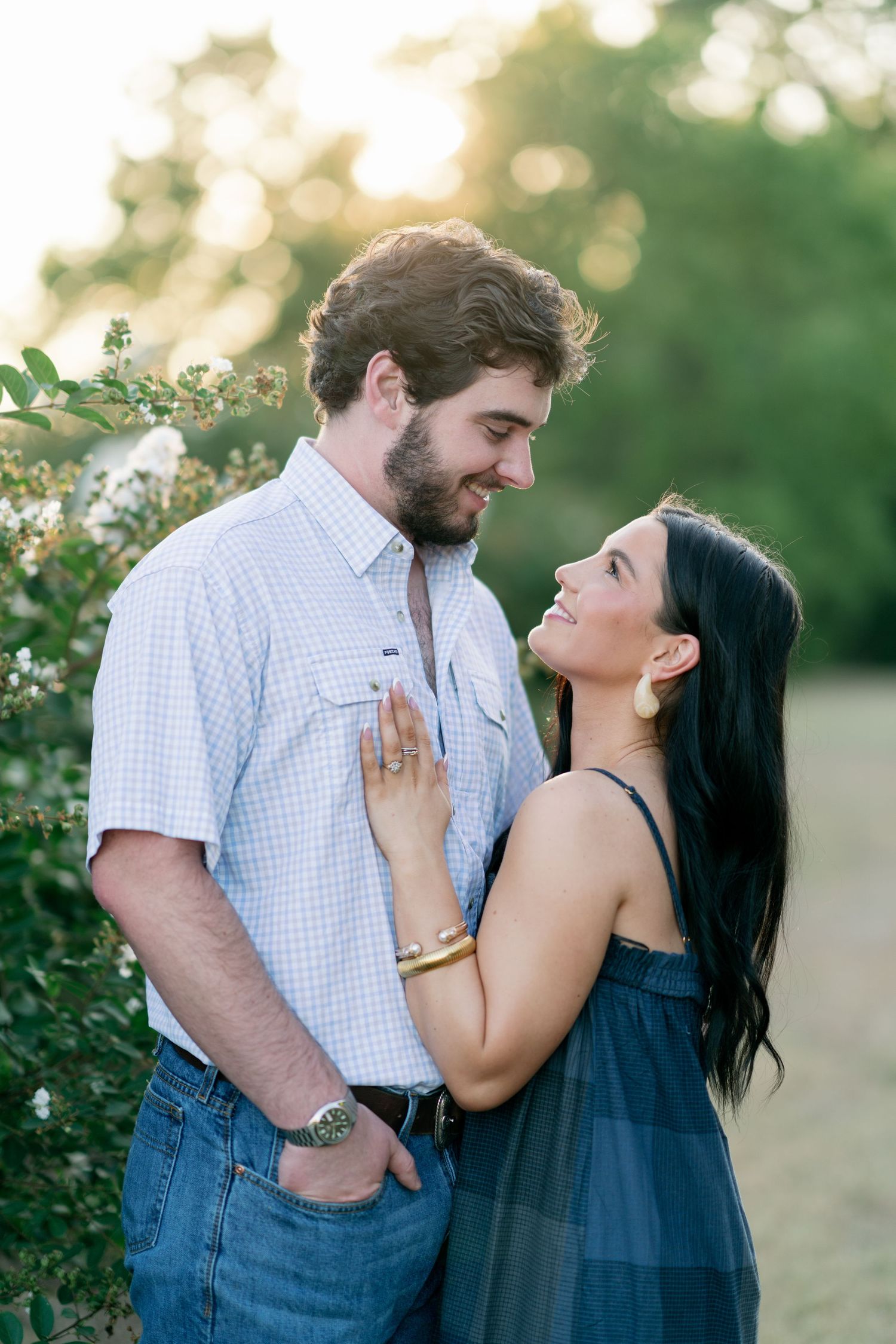 A couple shares a romantic moment outdoors at golden hour, woman in blue dress smiling up at bearded man in plaid shirt.